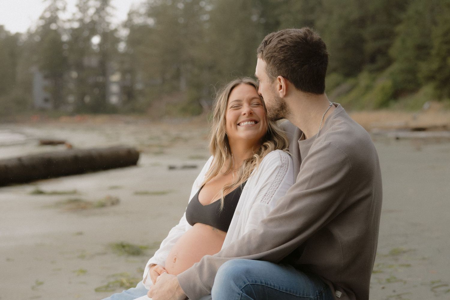Couple embraces on a misty Pacific Northwest beach with evergreen trees in background.