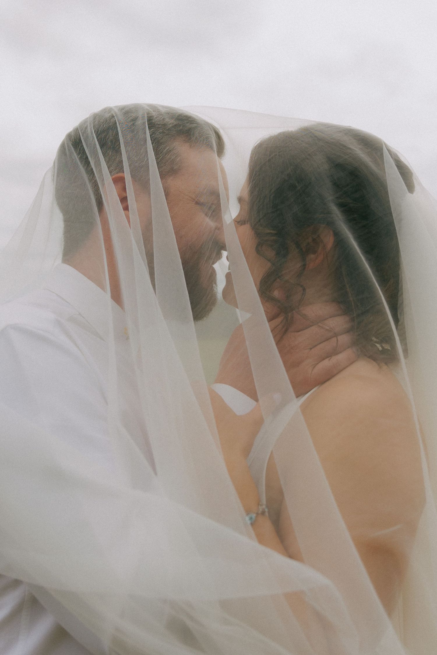 A bride and groom share an intimate moment under a flowing white veil, nearly kissing in a romantic wedding portrait.