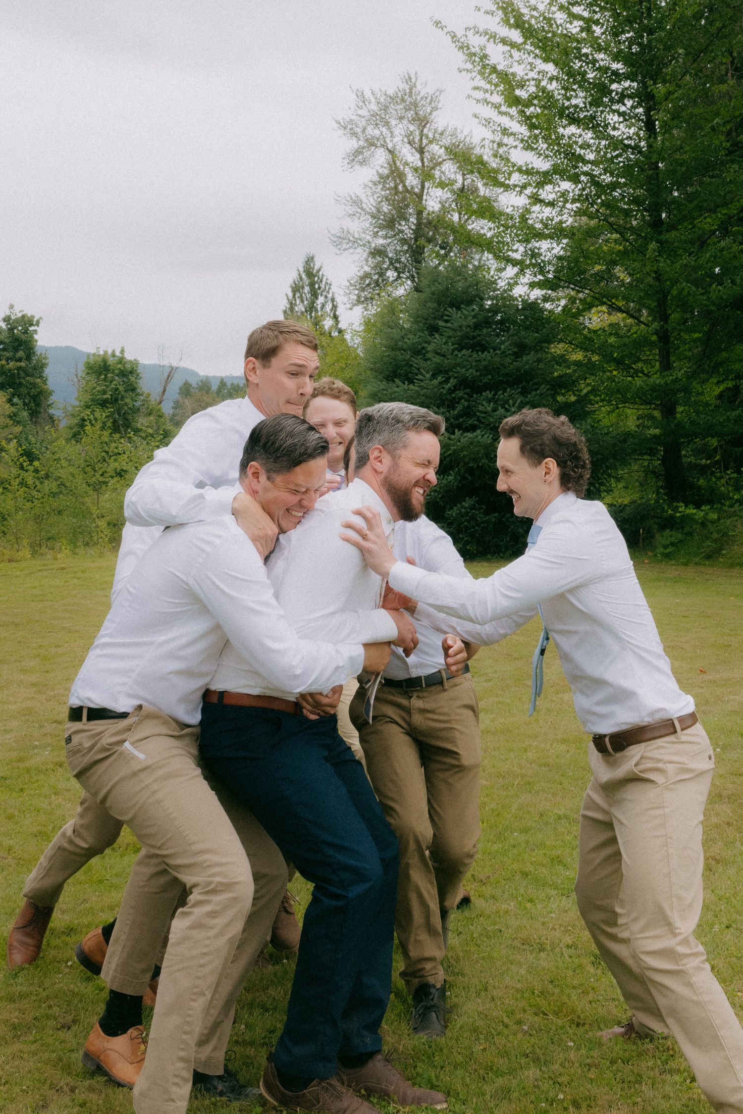 Groomsmen playfully tackle the groom outdoors at a wedding, dressed in white shirts and khaki pants.