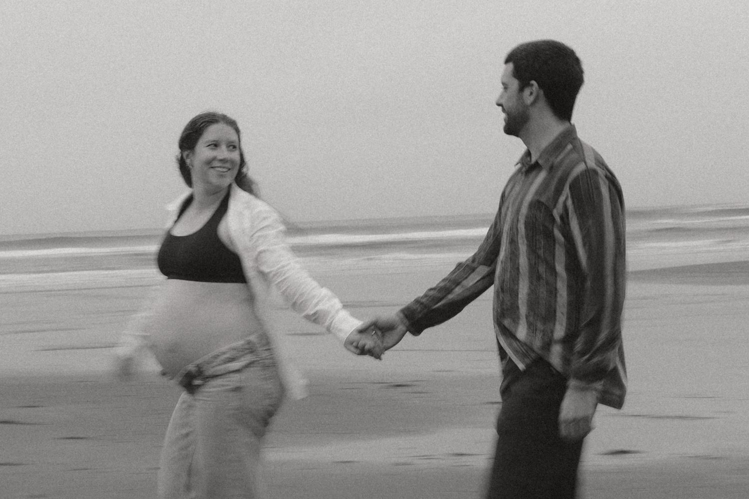 A couple holding hands on a beach, with the woman wearing white and visibly pregnant.