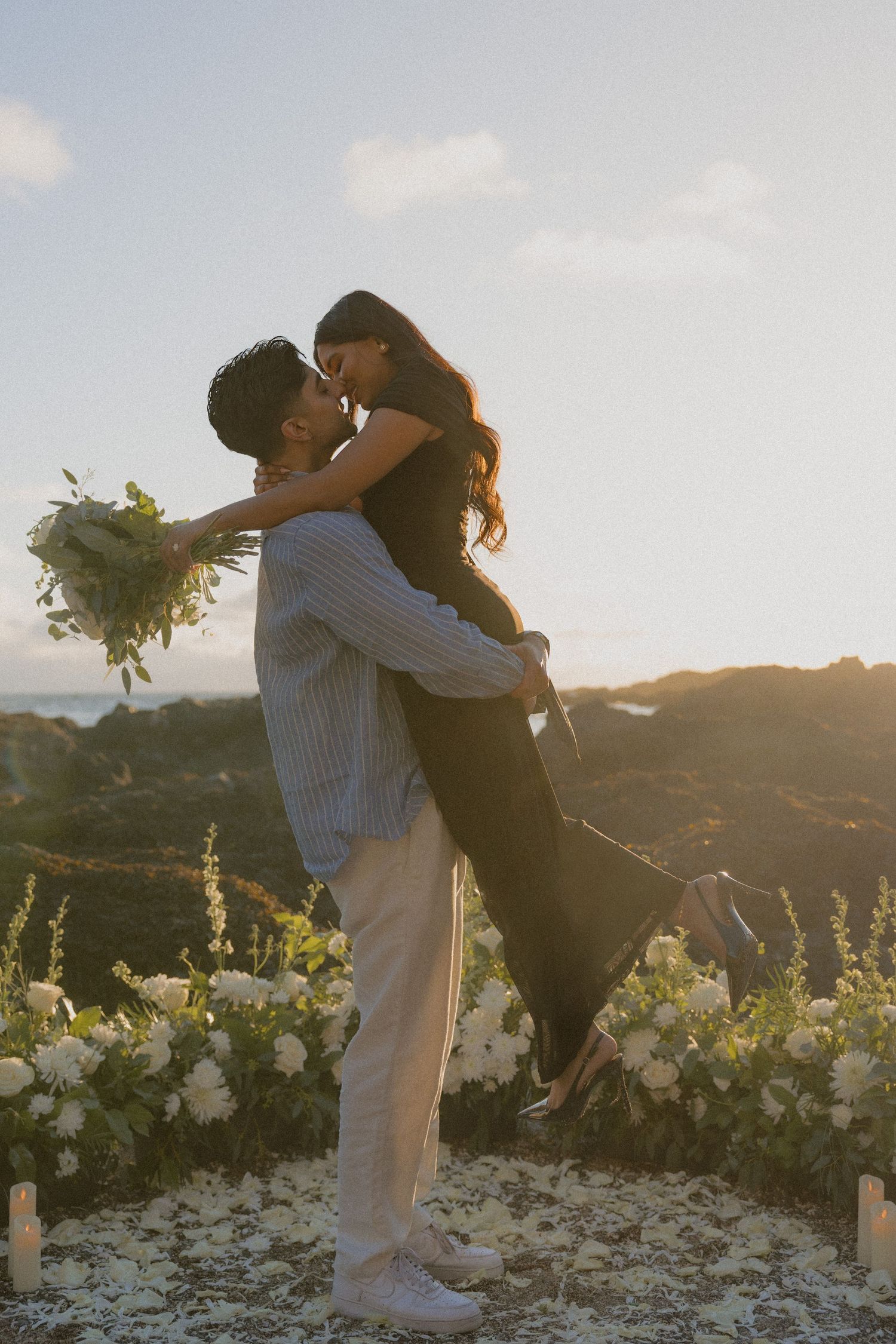 Romantic couple kissing at sunset with white floral arrangement during outdoor proposal.
