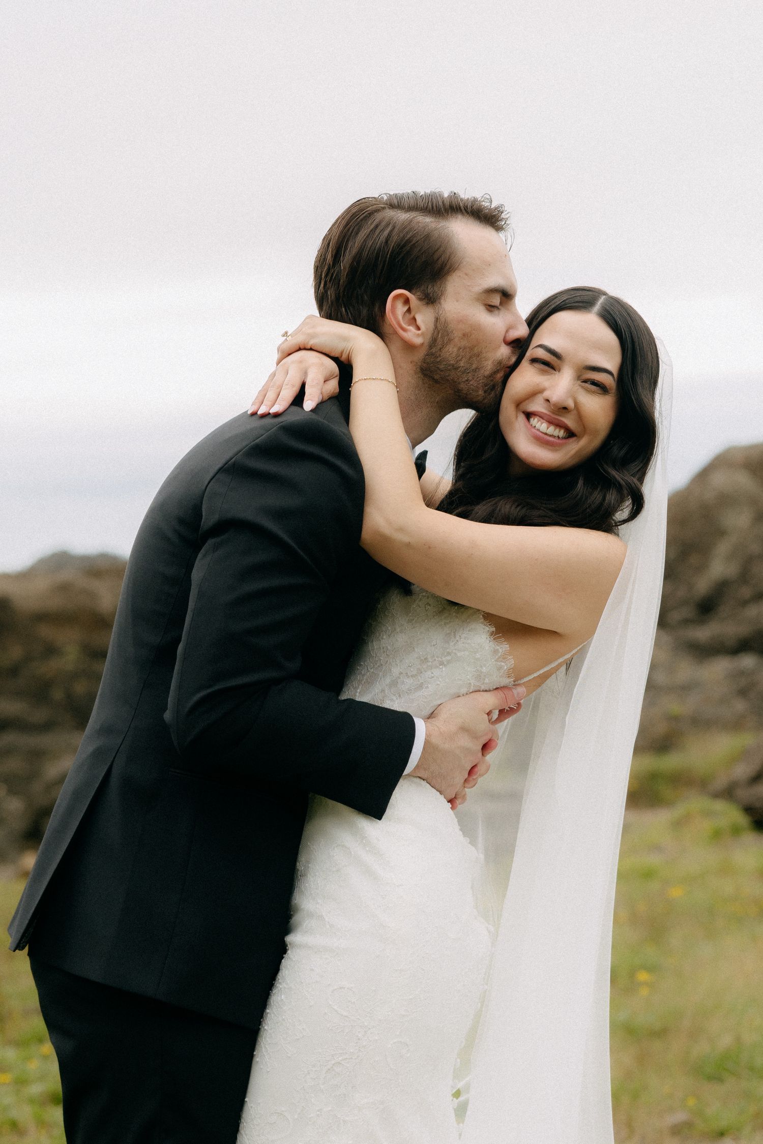 Groom in black suit kisses bride in white lace gown and veil outdoors on their wedding day.