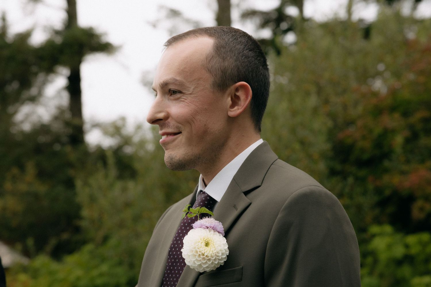 Smiling groom in gray suit with white boutonniere at outdoor wedding ceremony surrounded by greenery.