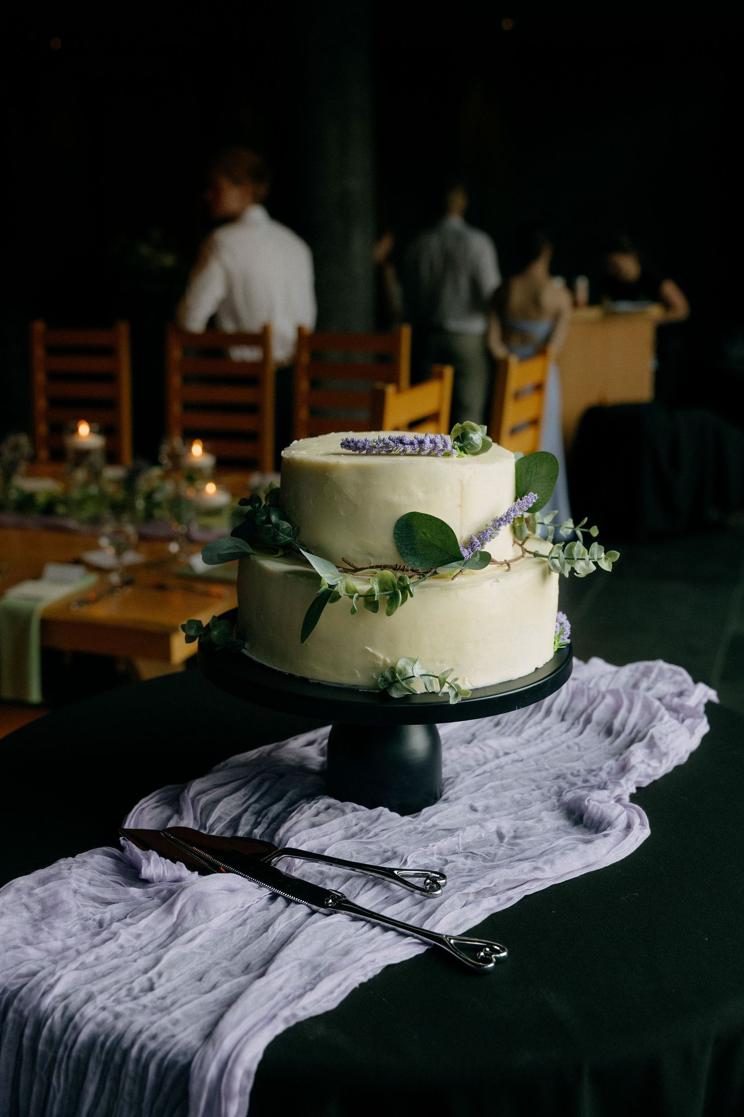 Elegant two-tier white wedding cake decorated with greenery and purple flowers on a black cake stand.