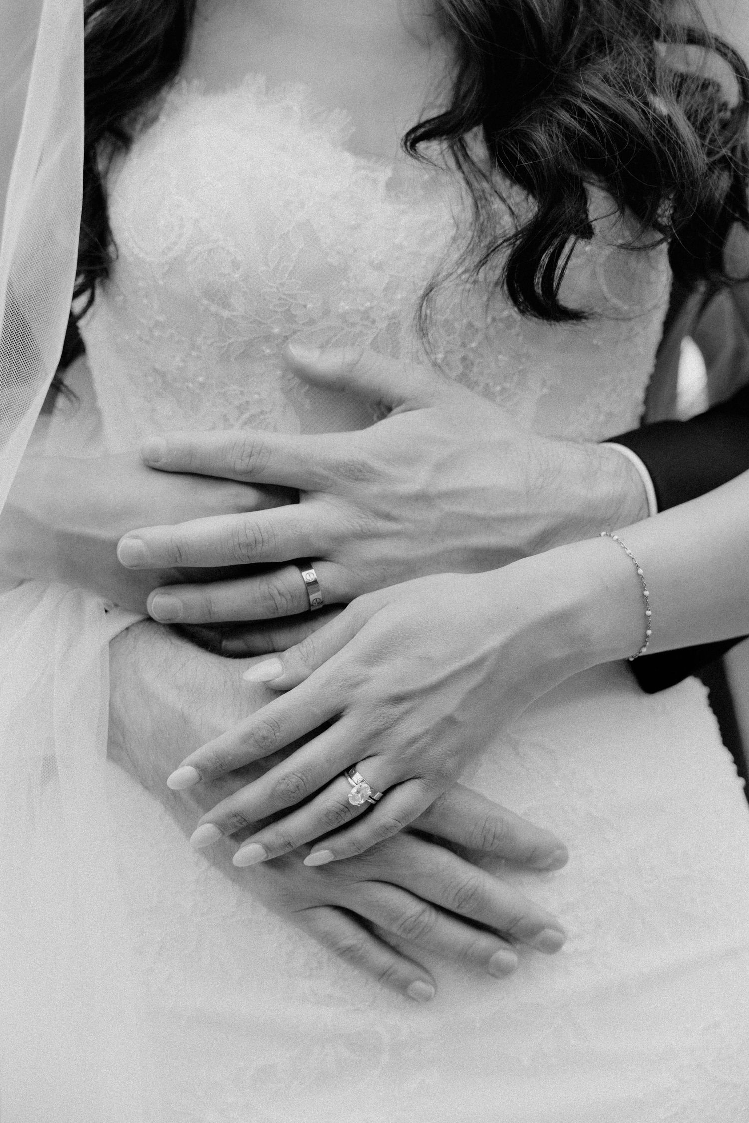 Black and white photo of bride and groom hands showing wedding rings, bride in lace dress with veil.