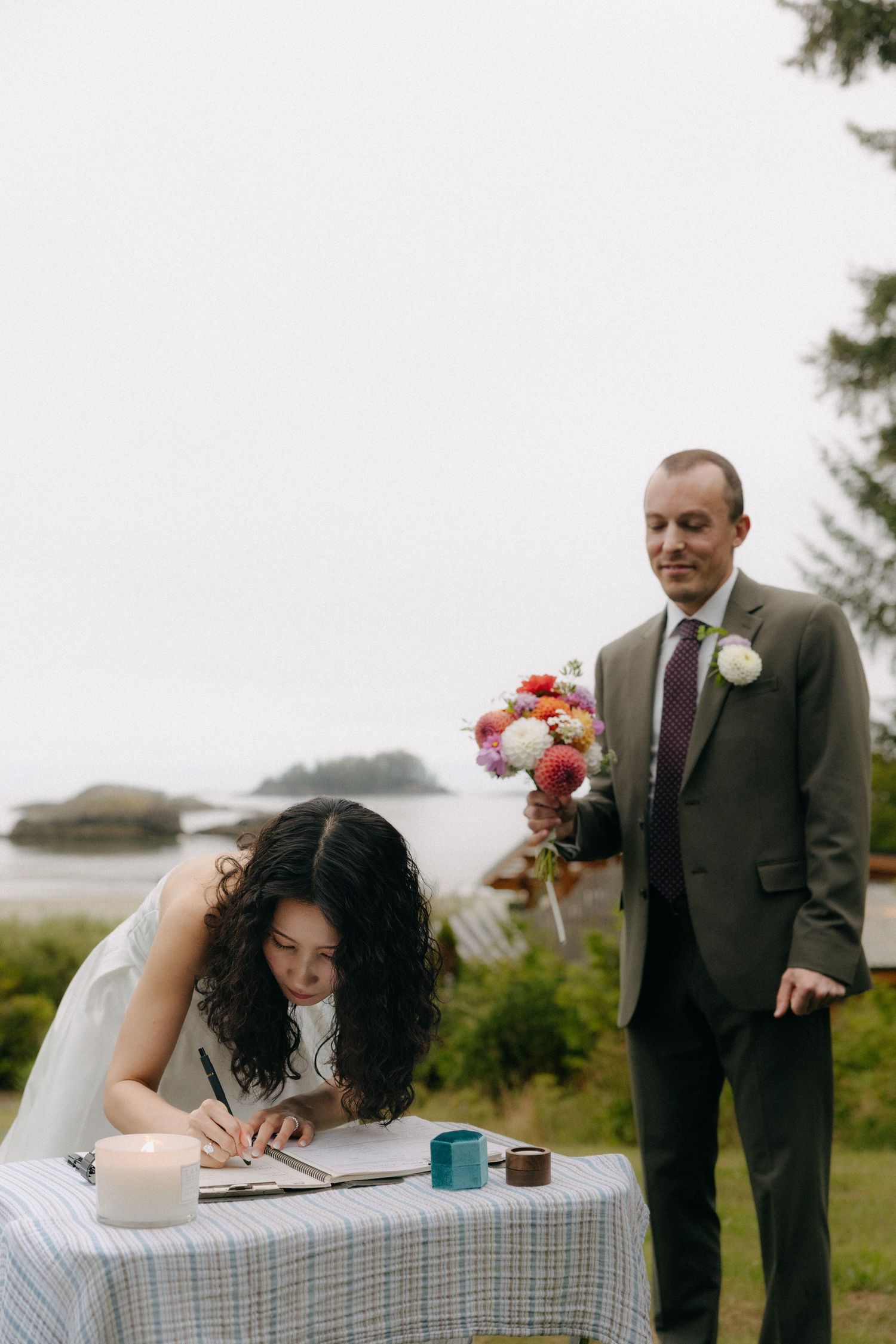 Bride signing wedding documents at outdoor table while groom holds colorful bouquet near scenic coastal backdrop.