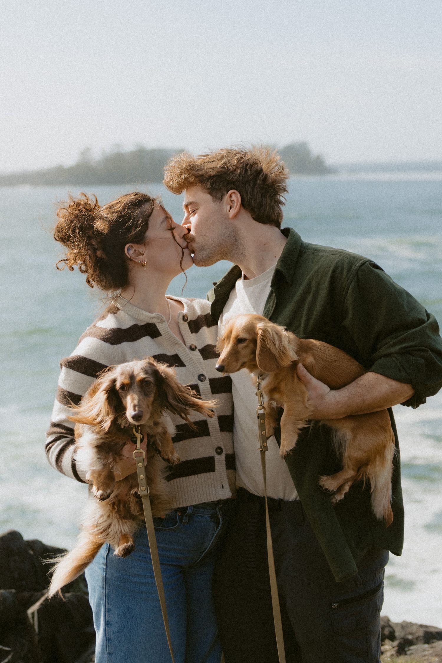 Engaged couple kissing while holding two golden dachshund dogs on rocky ocean coastline.