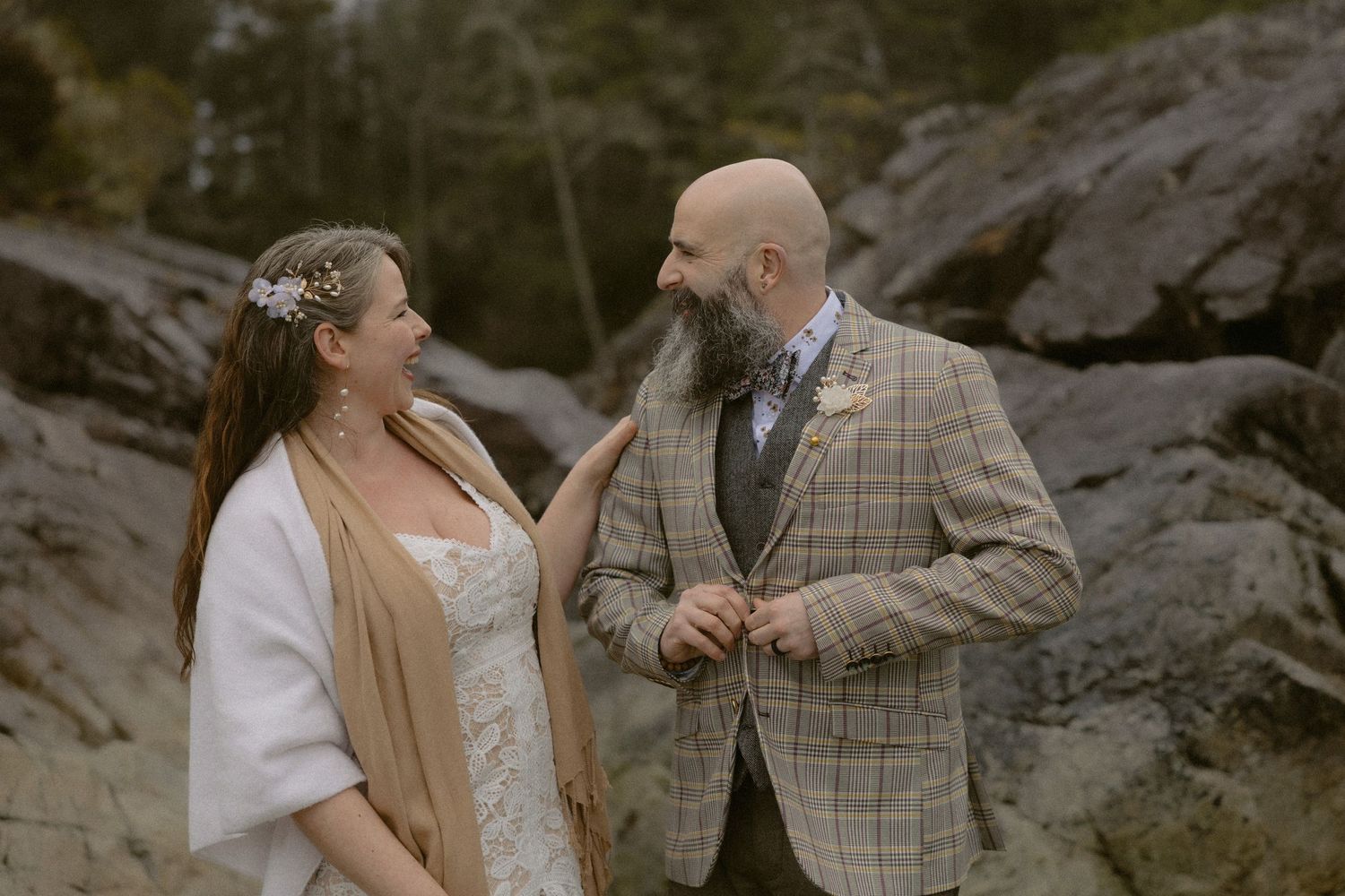 A couple shares an intimate moment against rocky cliffs wearing wedding attire and plaid suit.