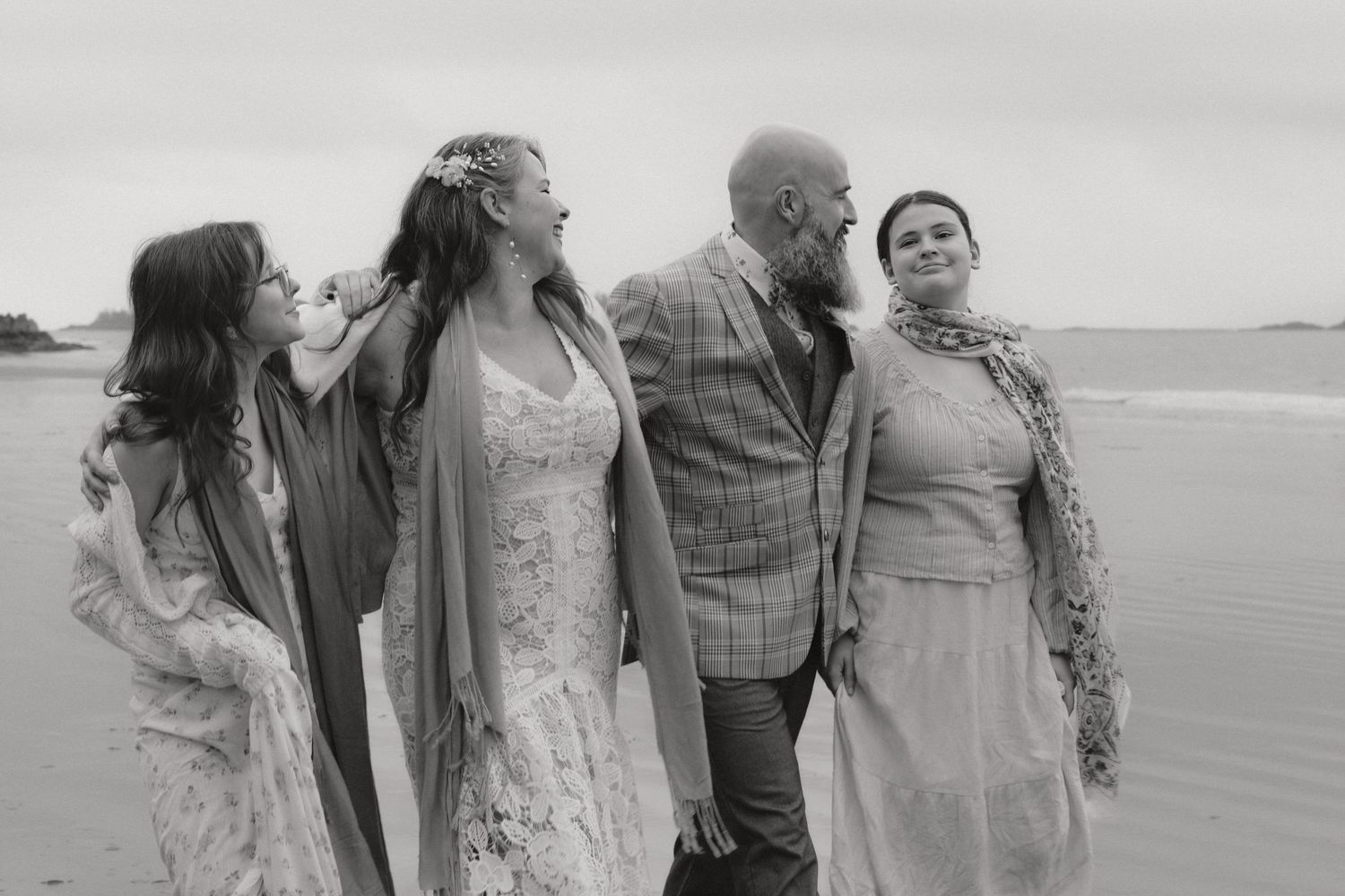 A wedding party laughs together while standing on a beach in black and white.