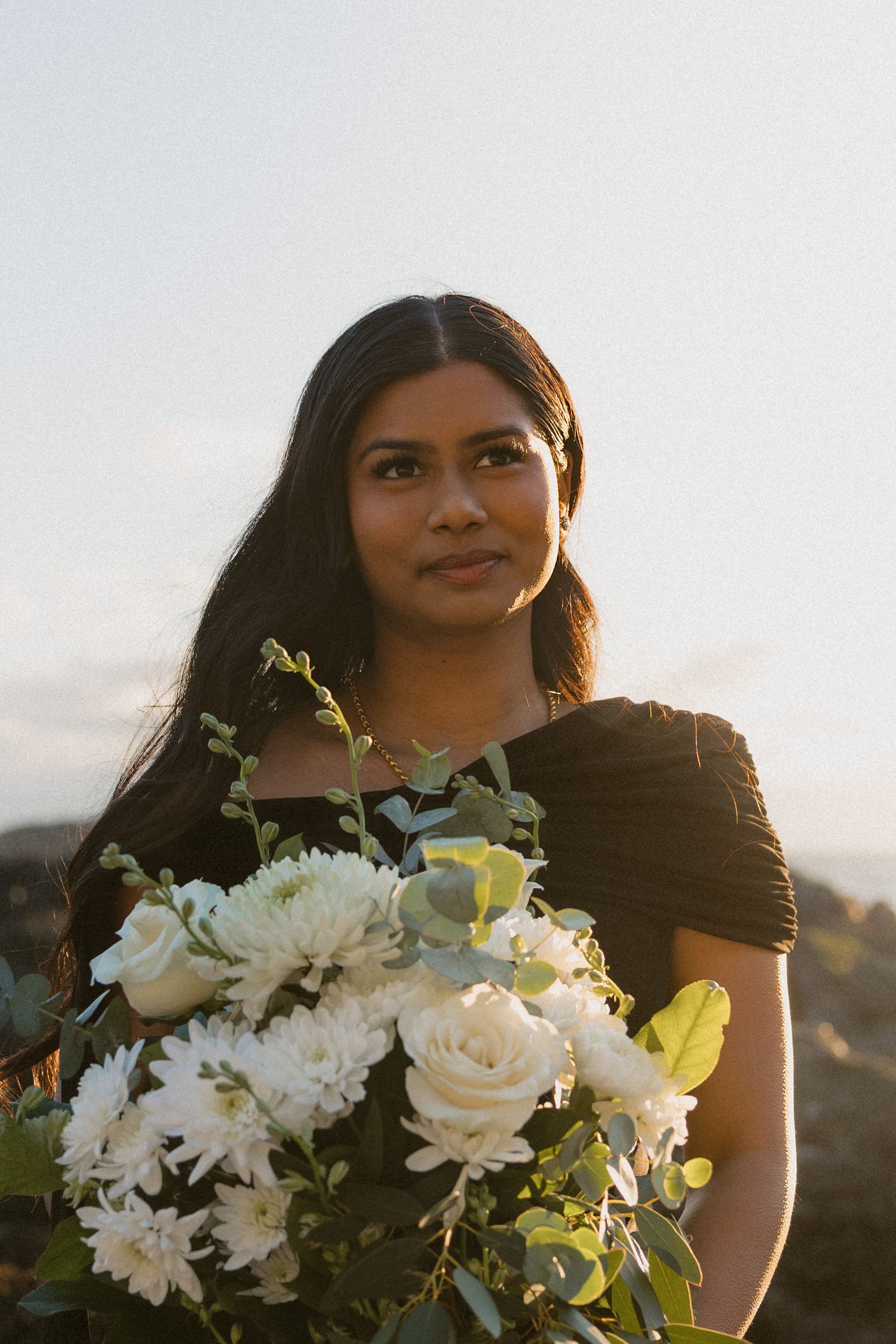 Young woman in black dress holding elegant white floral bouquet outdoors at sunset.
