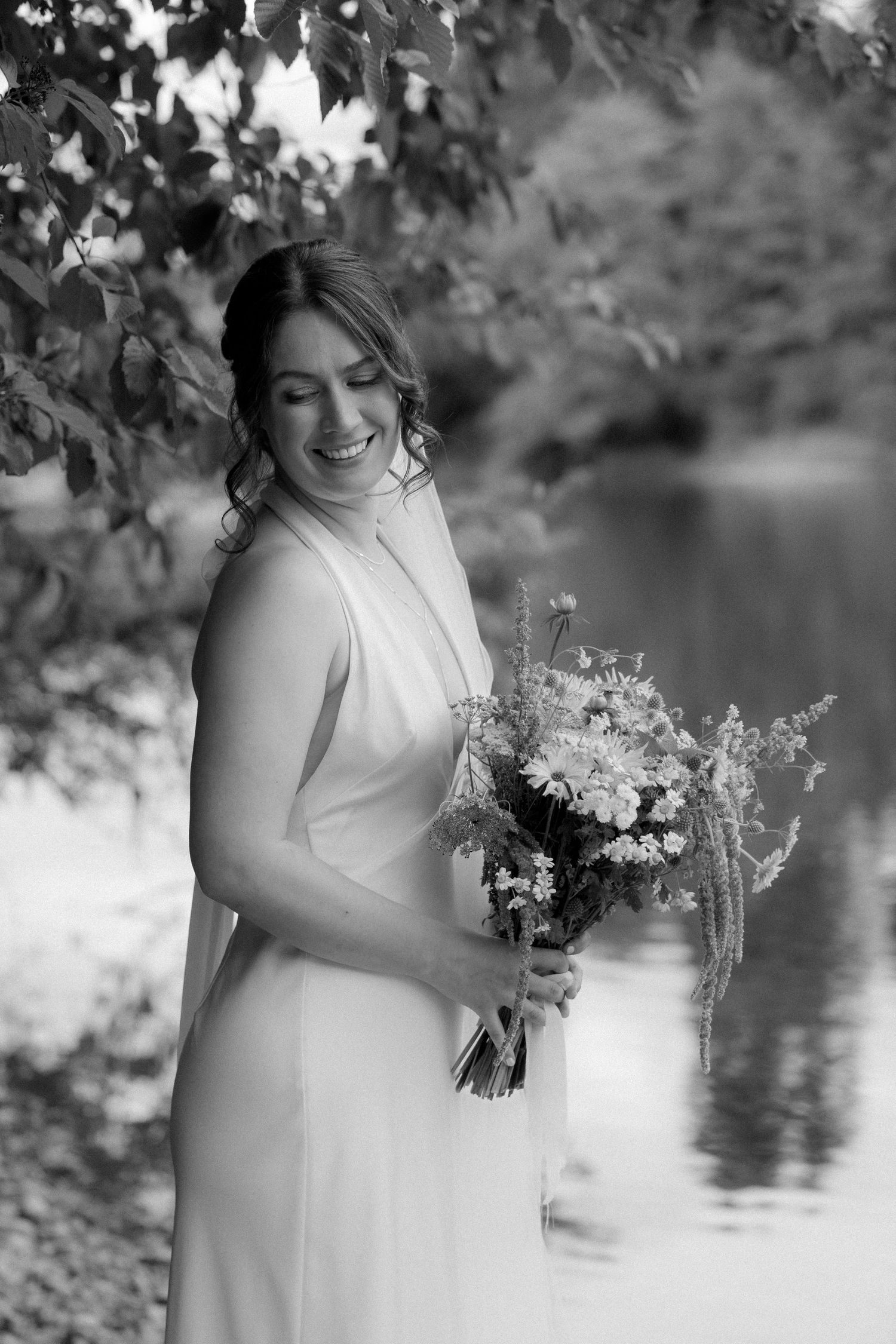 Black and white photo of a smiling bride holding a wildflower bouquet near a reflective lake.