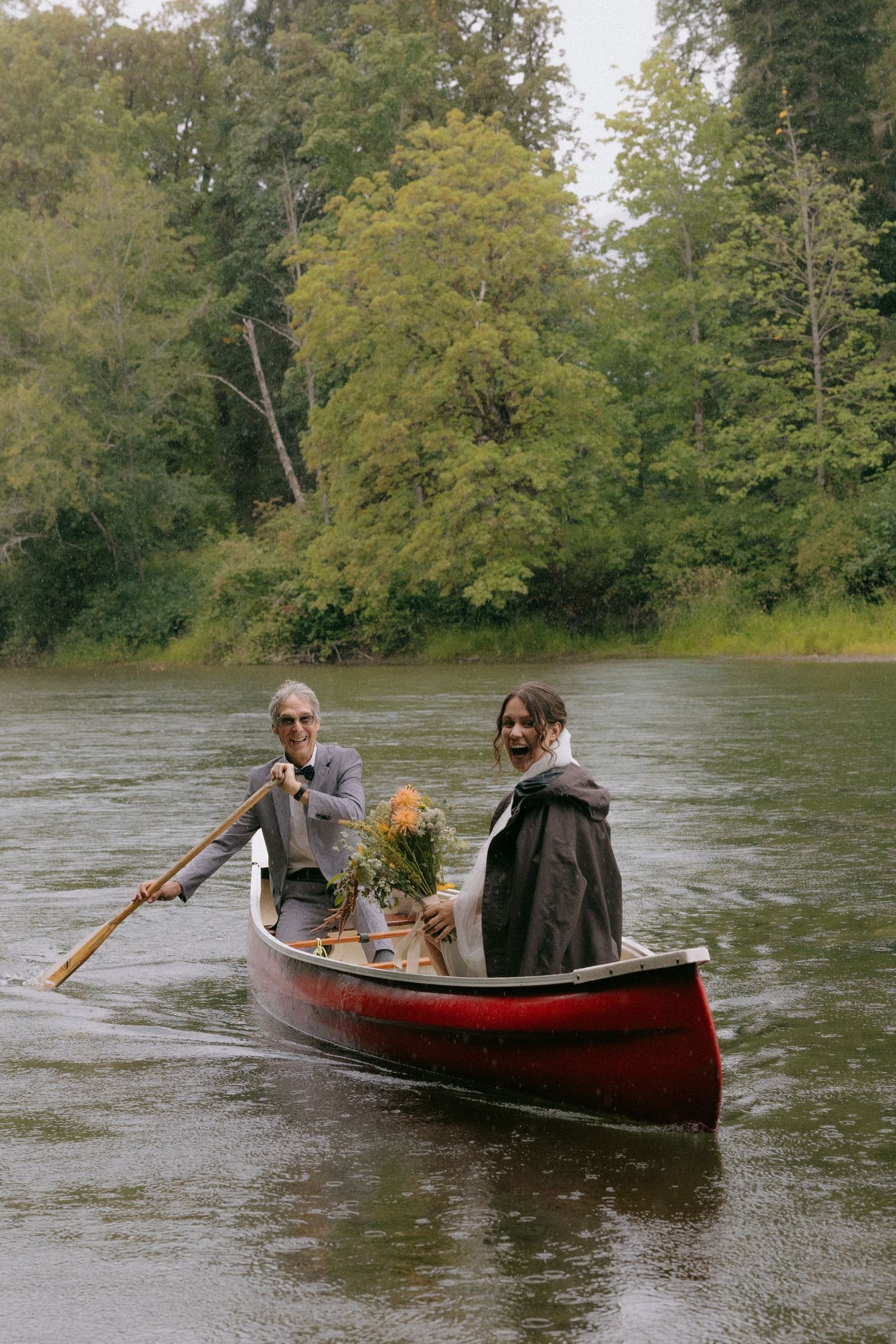 Couple in a red canoe on a river, one paddling, the other holding a bouquet, surrounded by lush green trees.