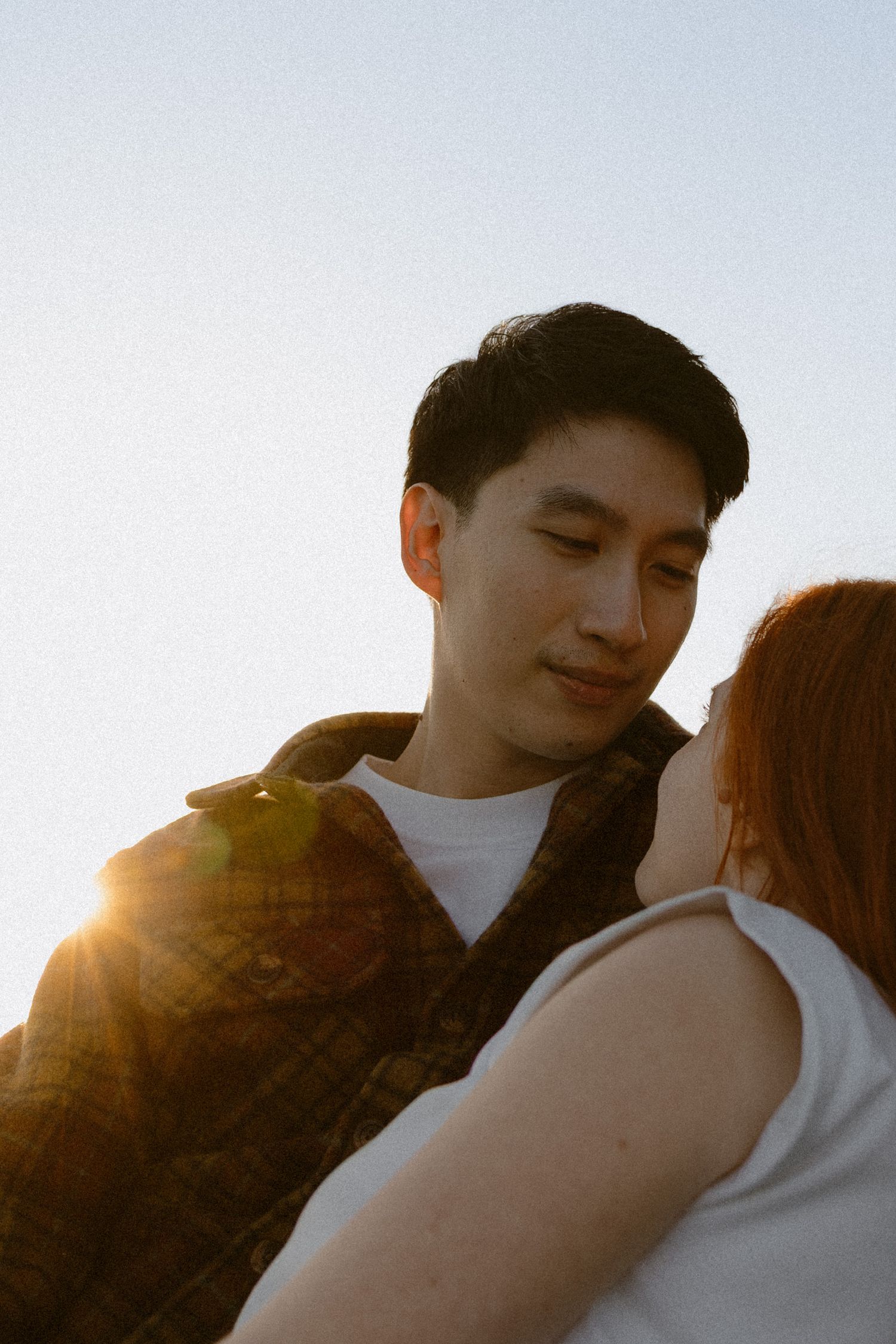 A couple shares a tender moment at golden hour, with warm sunlight glowing behind them against a clear sky.
