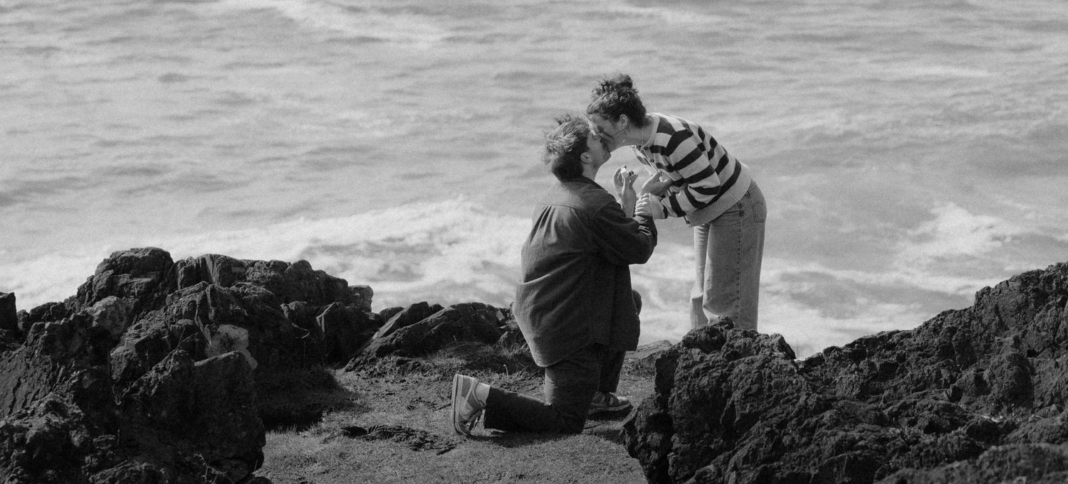 Black and white photo of couple sharing a romantic proposal moment on rocky coastal cliffs by the sea.