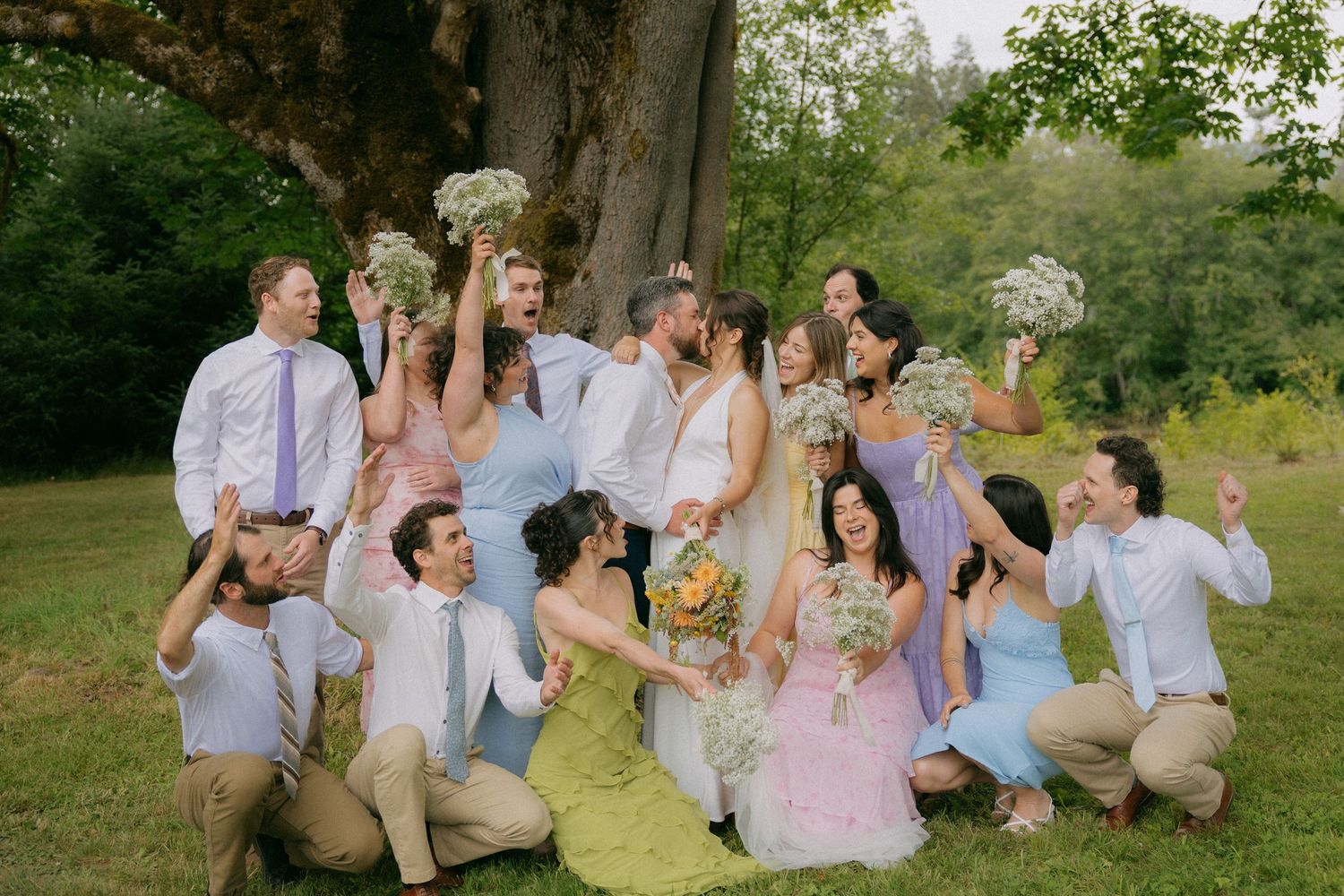 Wedding party celebrating outdoors under large tree, colorful dresses, bouquets raised joyfully.