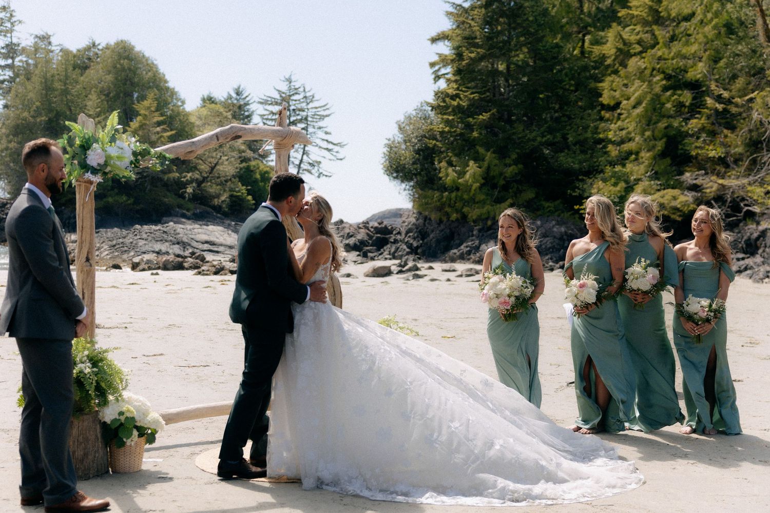 Bride and groom kiss at outdoor beach wedding ceremony with bridesmaids in sage green dresses and forest backdrop.
