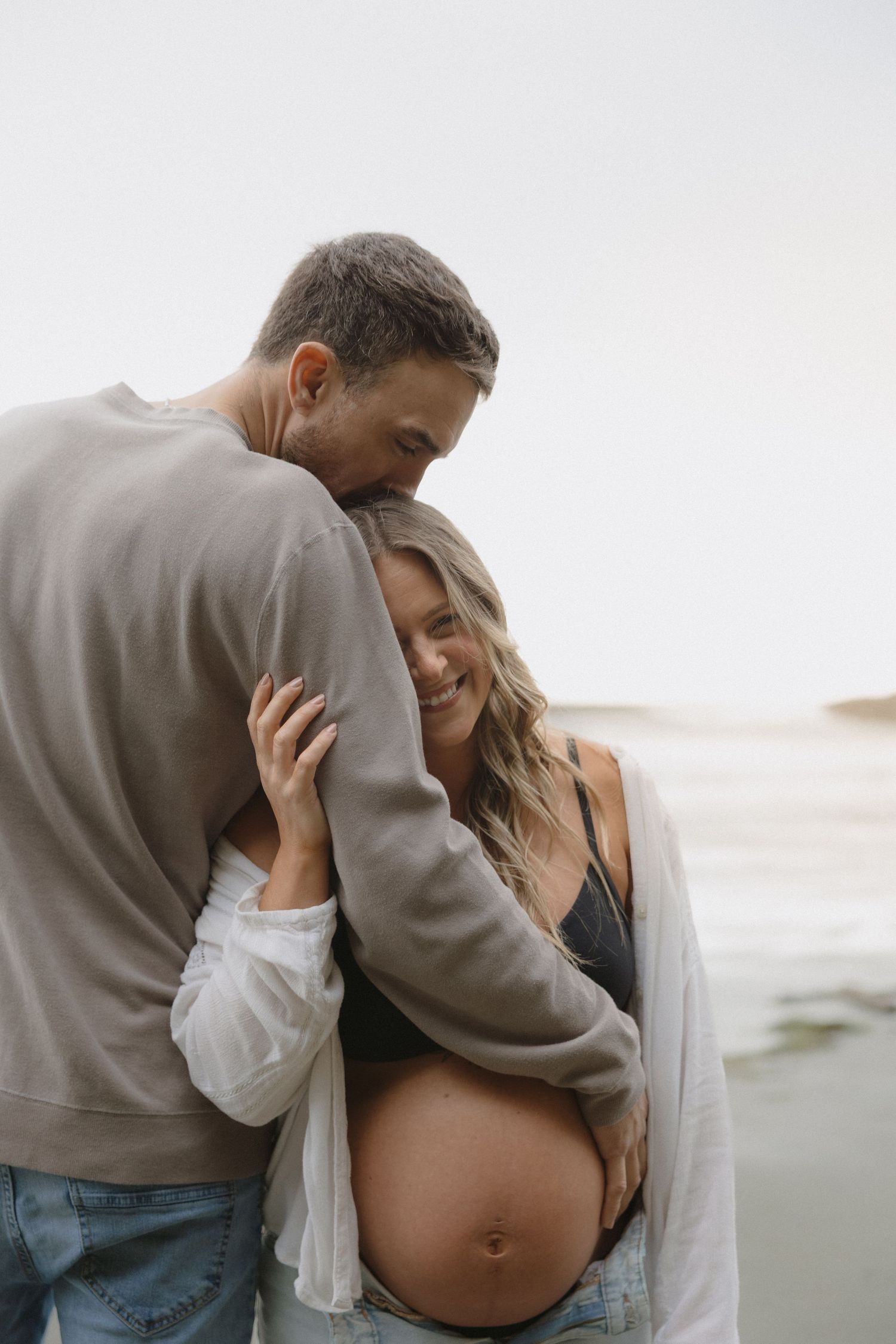 Tender embrace between couple during maternity photoshoot on overcast beach.