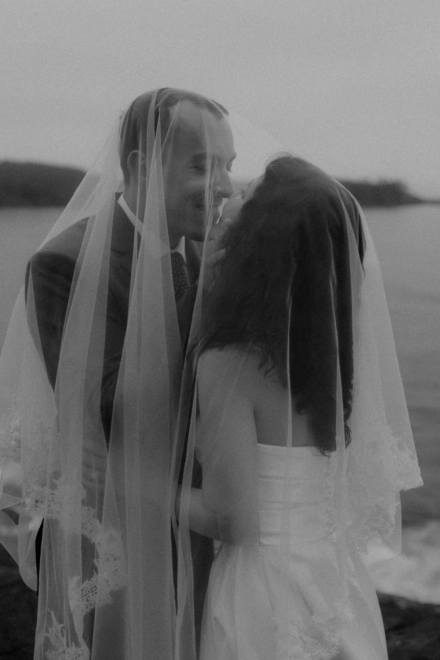 Black and white photo of a bride and groom sharing a romantic moment under a lace wedding veil by the ocean.