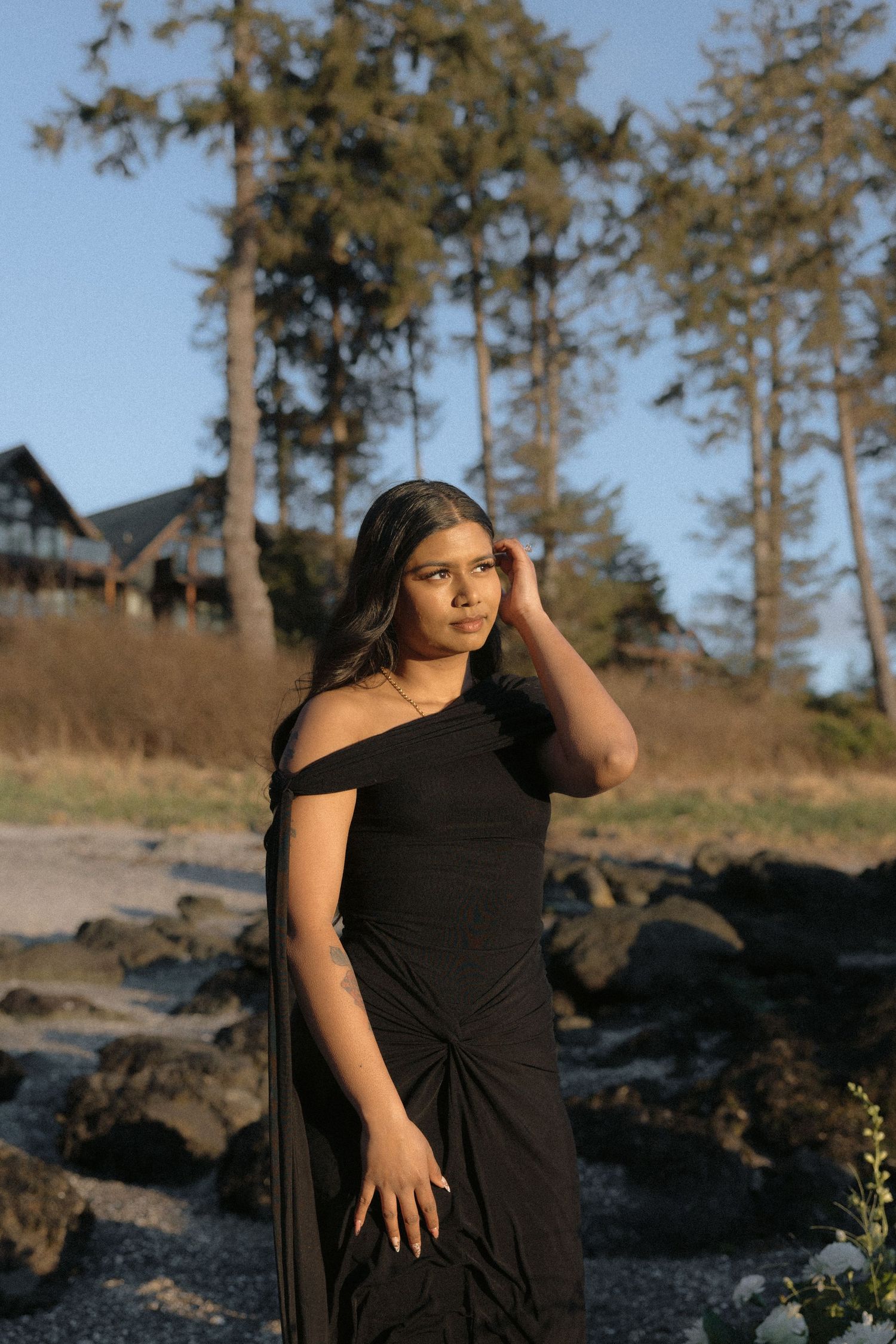 Young woman in black off-shoulder dress posing on rocky beach at sunset.