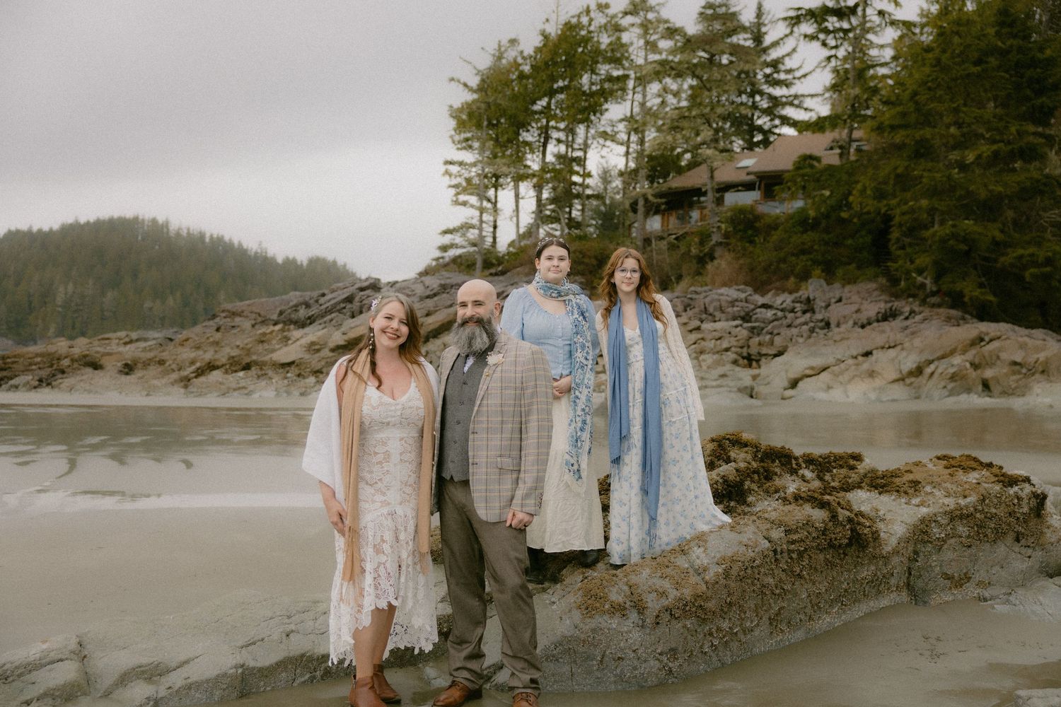 Wedding party stands on rocky beach with evergreen trees and cloudy sky in Pacific Northwest setting.