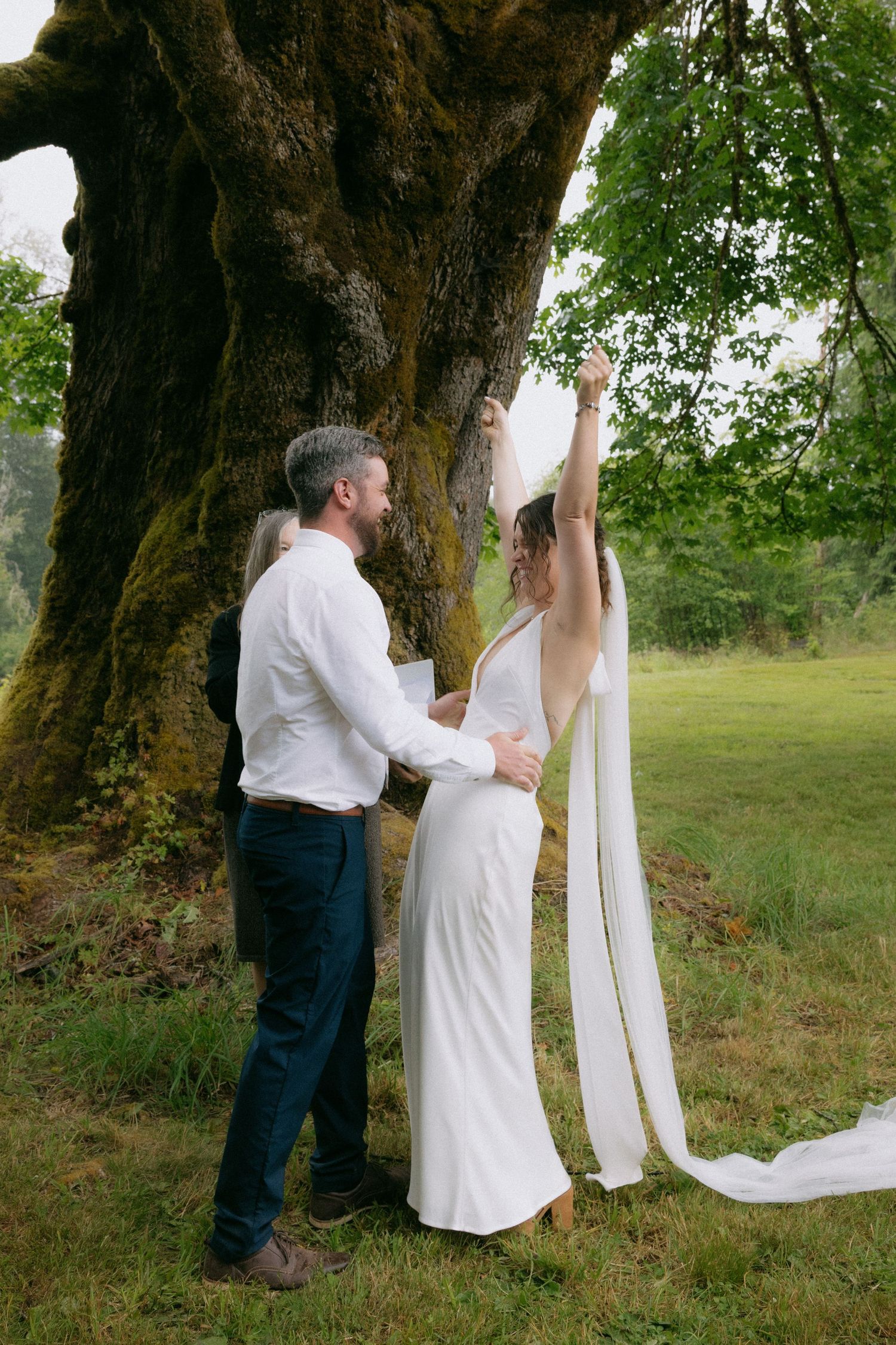 Bride in white gown raises veil beside groom near massive moss-covered tree in lush green setting.