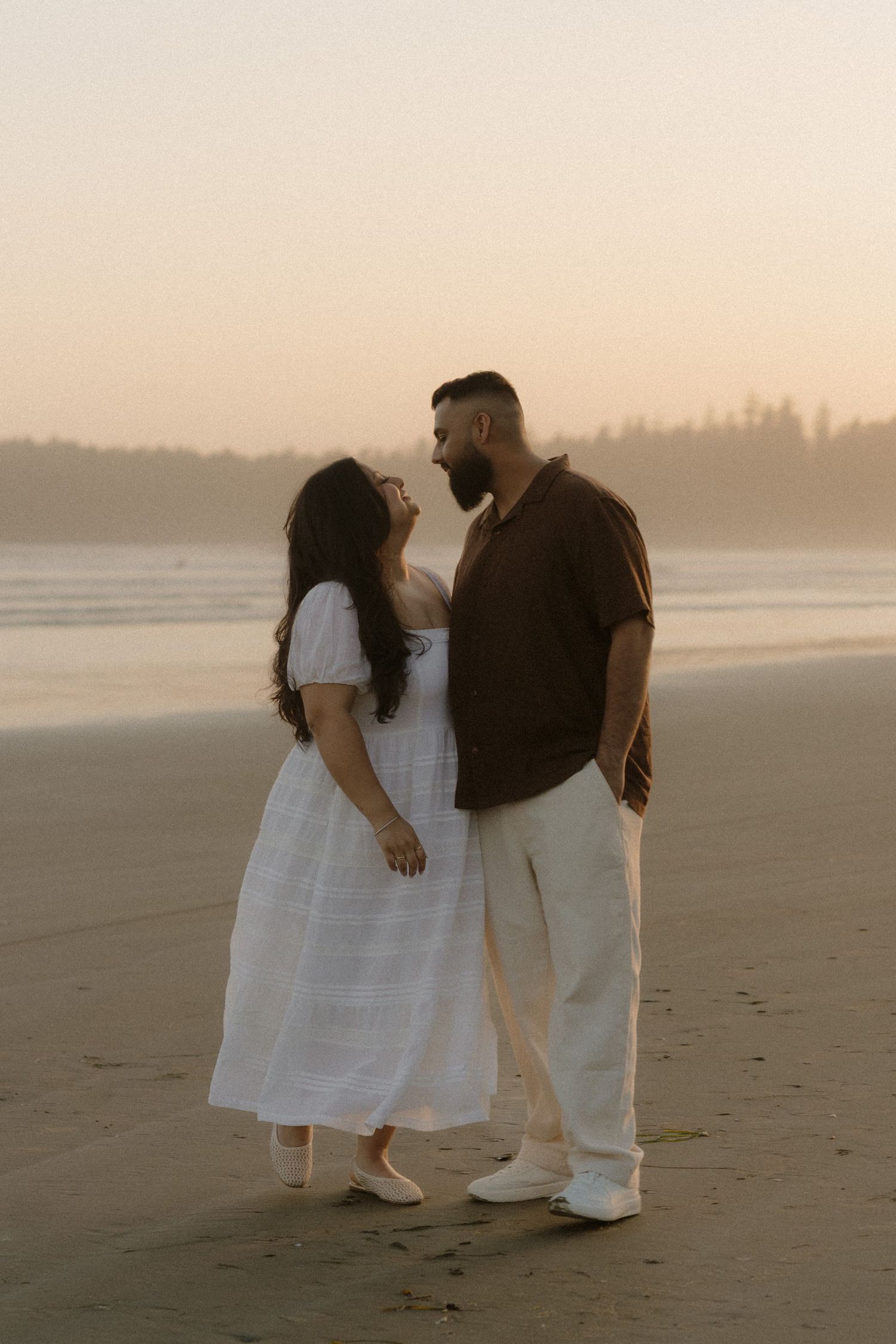 Couple sharing a romantic moment on a misty beach at sunset, woman in white dress, man in dark shirt.