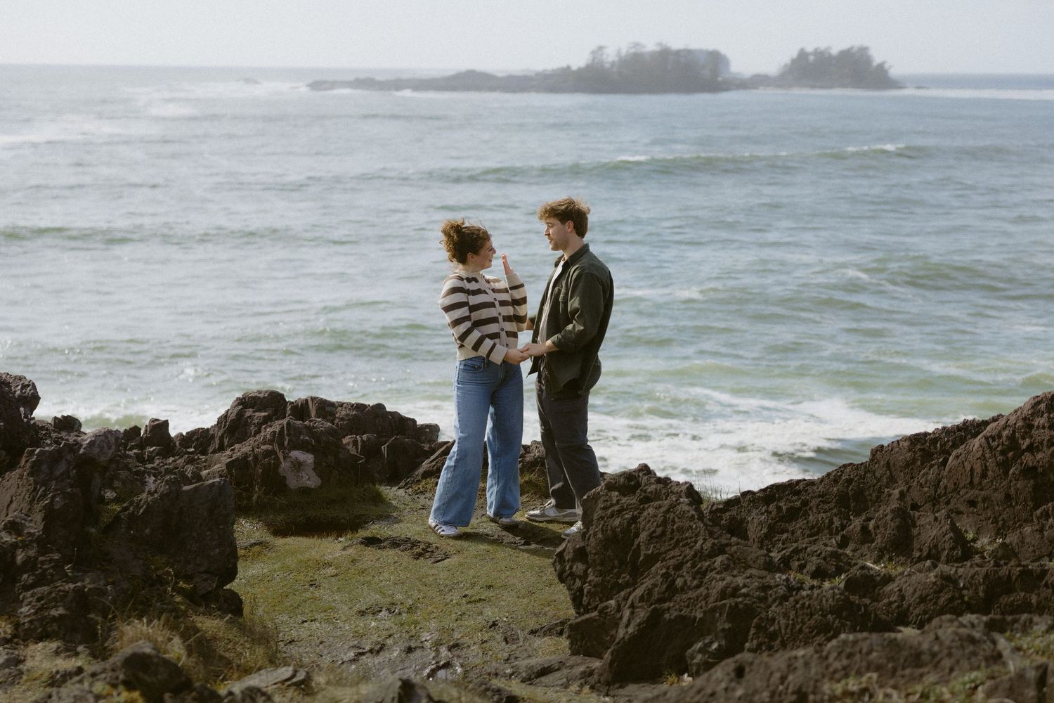 Couple holding hands on rocky ocean shore with waves and island in background.