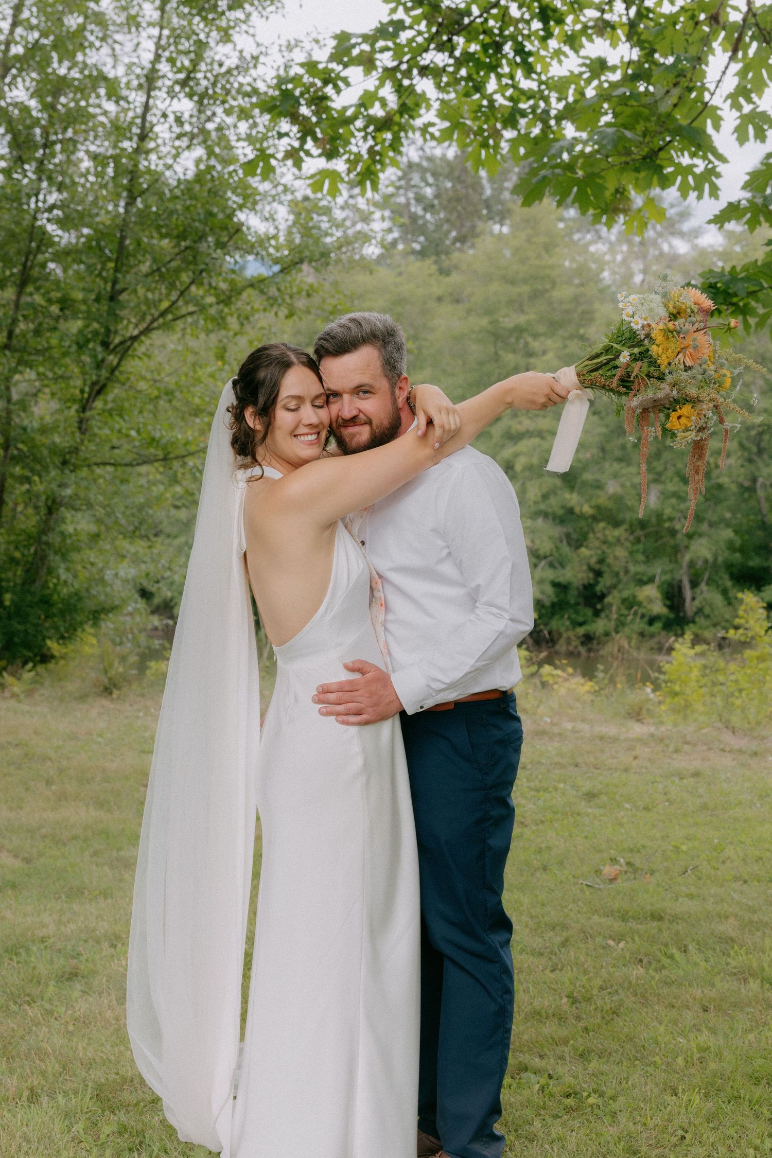 Bride and groom embrace outdoors in lush green setting, bride in backless white gown with veil holding wildflower bouquet.