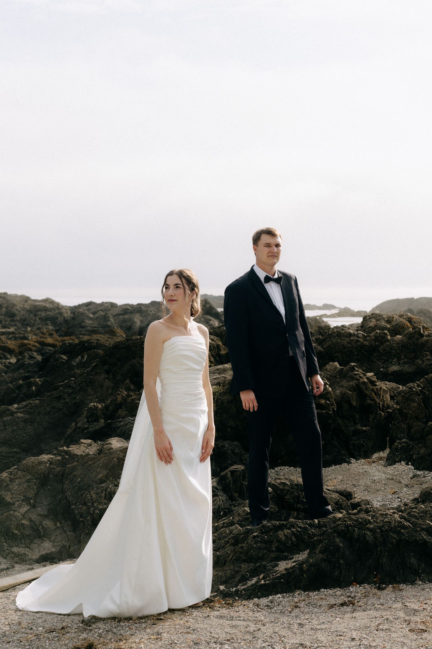 Bride in white strapless gown and groom in black tuxedo posing on rocky beach.