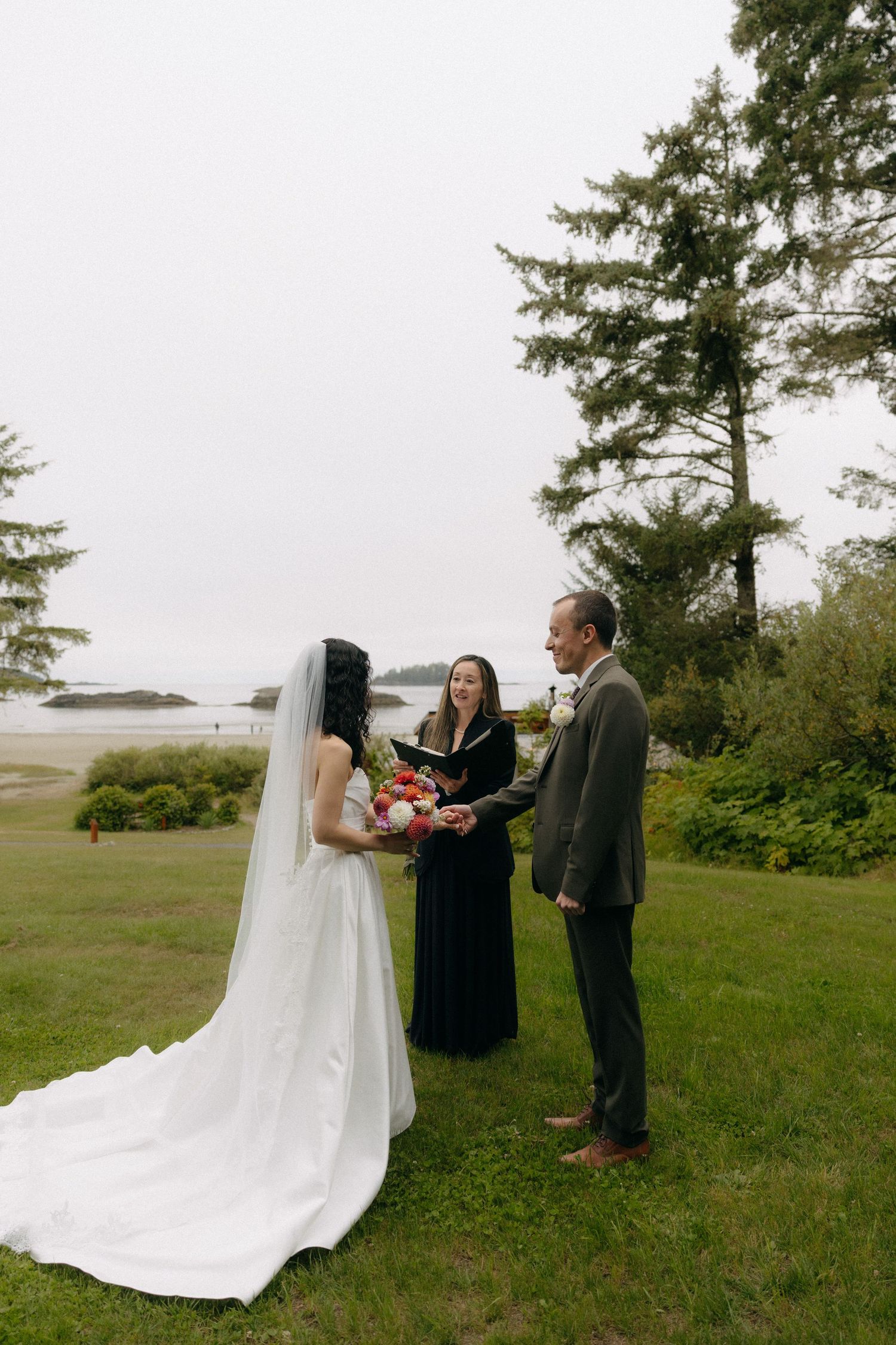 Bride and groom exchange vows outdoors with officiant, ocean view backdrop, lush greenery, bride holding floral bouquet in white gown.
