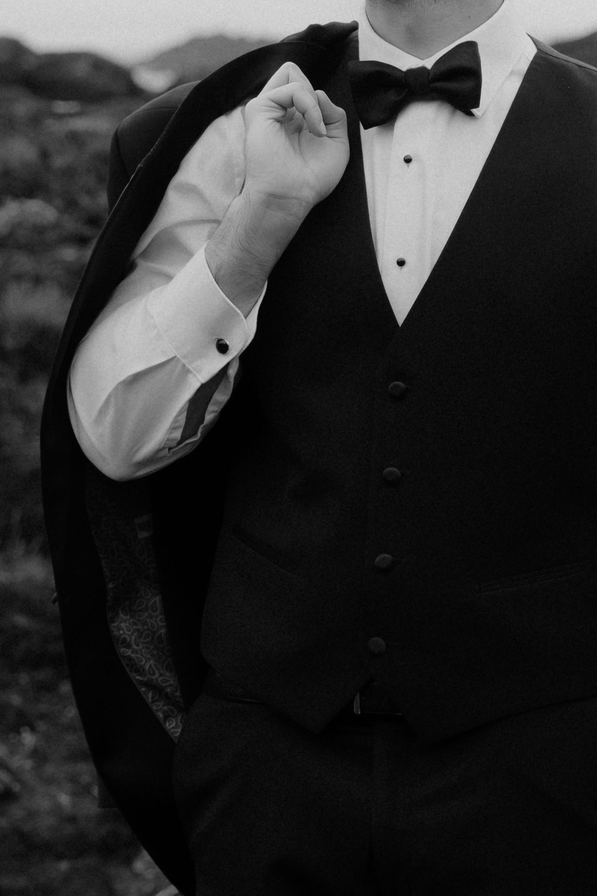 Groom in black tuxedo vest and bow tie holds jacket over shoulder in elegant black and white wedding photo.