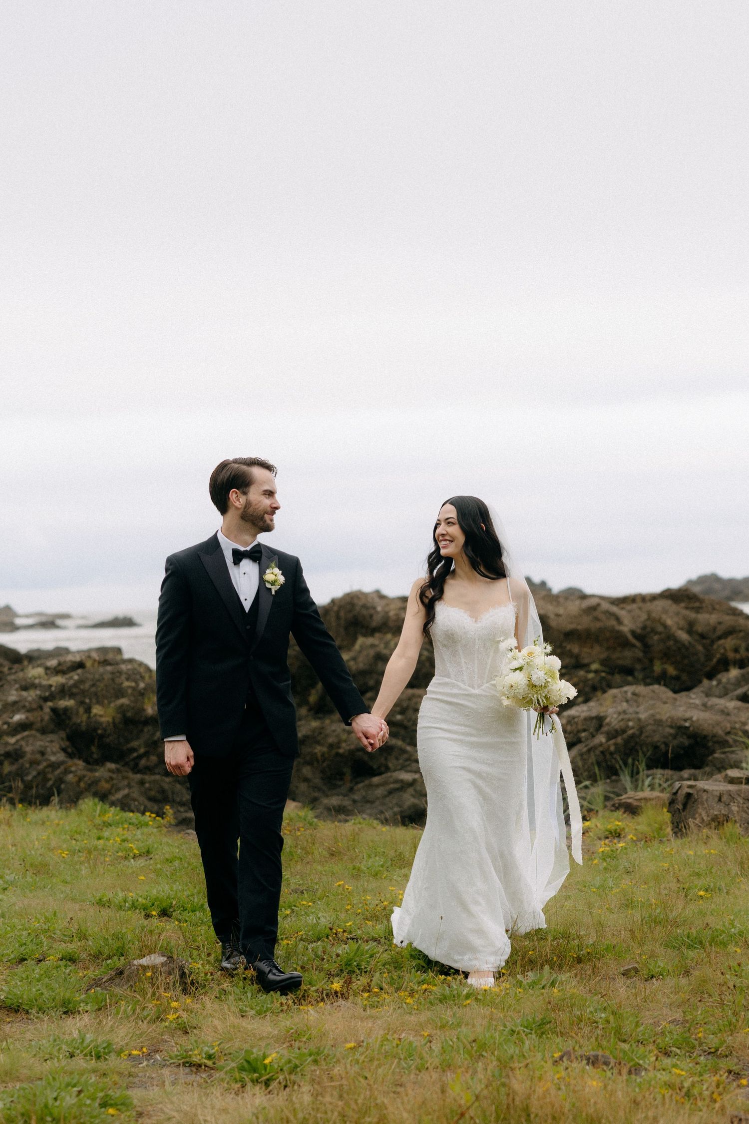 A bride and groom hold hands walking along a rocky coastal bluff, she in a white gown with bouquet, he in a black tuxedo.