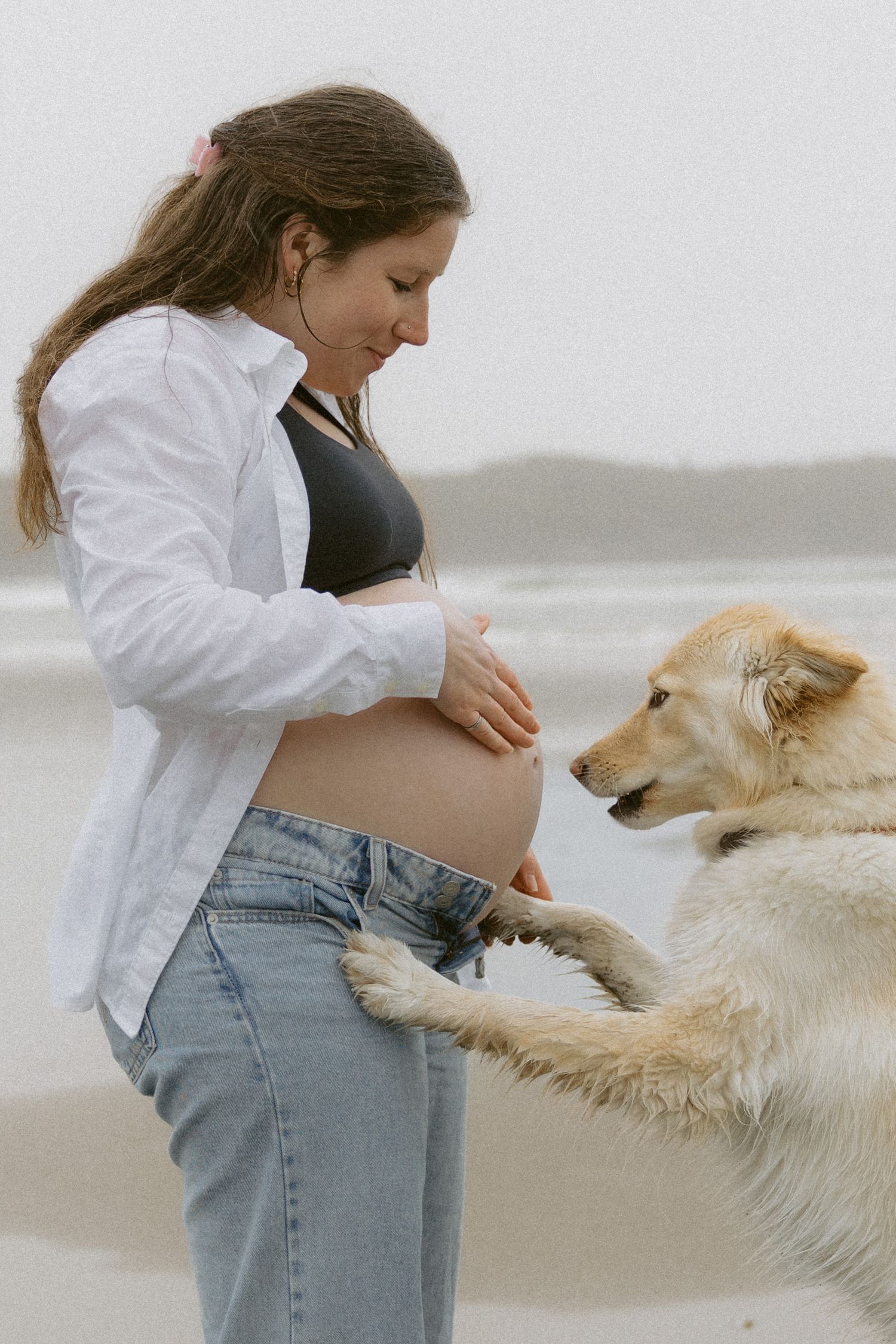 A woman in jeans and white shirt plays with a golden retriever dog on a misty beach.
