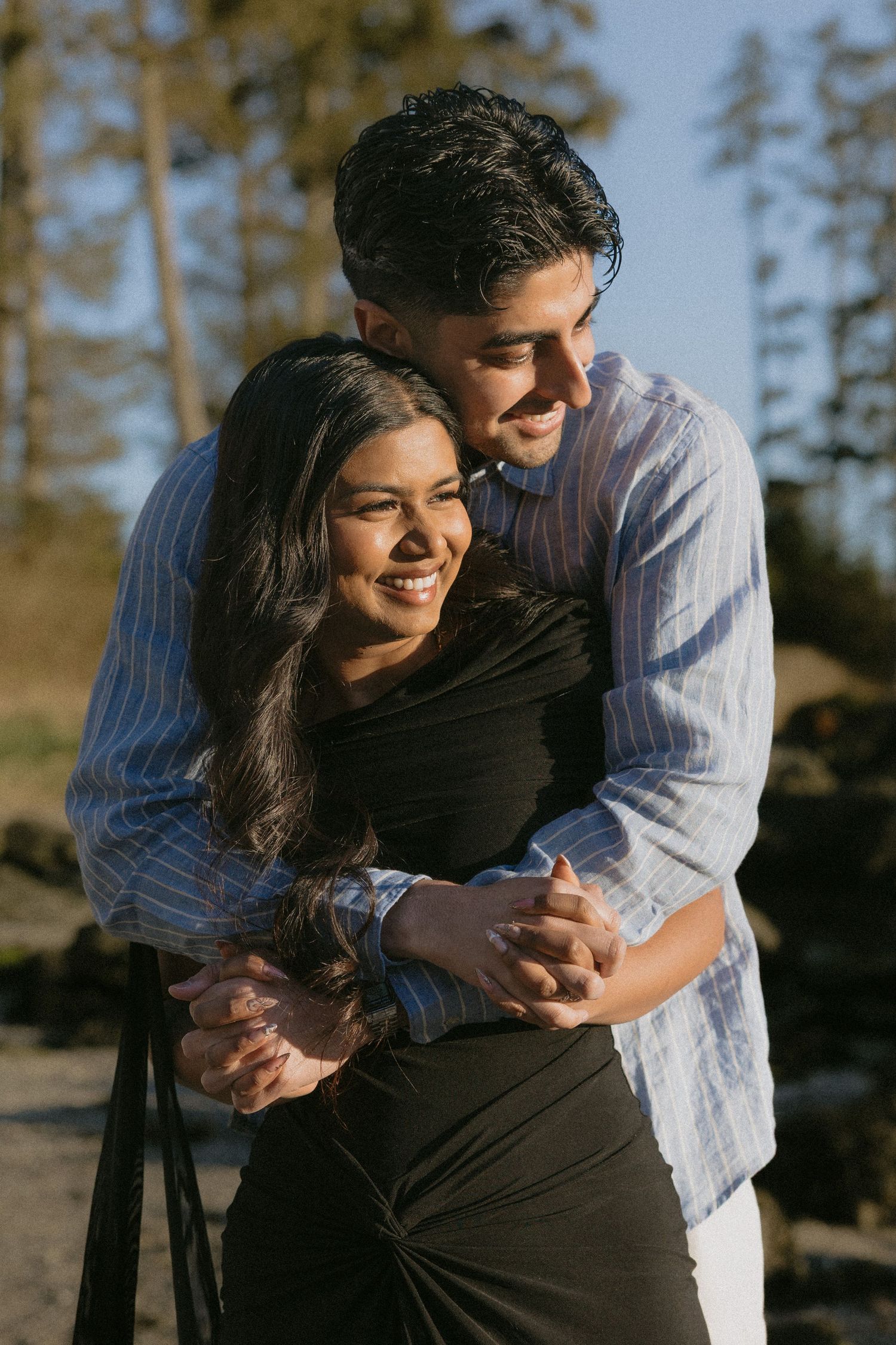 Happy couple embracing warmly, man hugging woman from behind, both smiling in sunny outdoor setting.