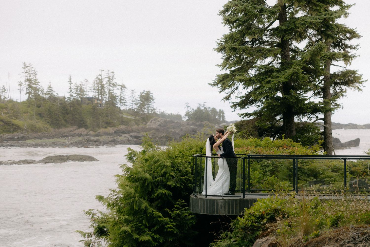 A couple shares a romantic kiss on a scenic overlook surrounded by lush trees and misty coastal waters.