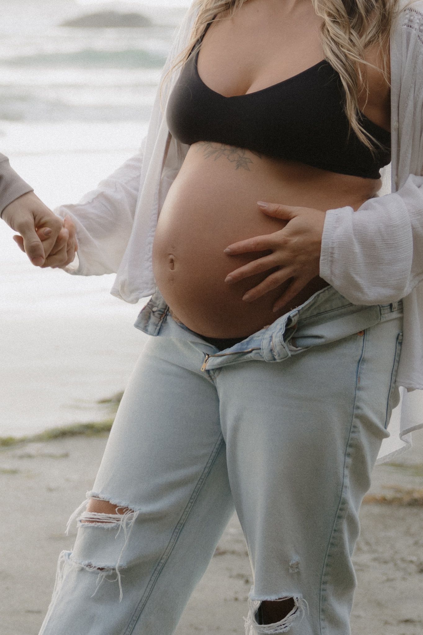 Intimate maternity photo session showing pregnant belly with light blue ripped jeans and black top on beach.