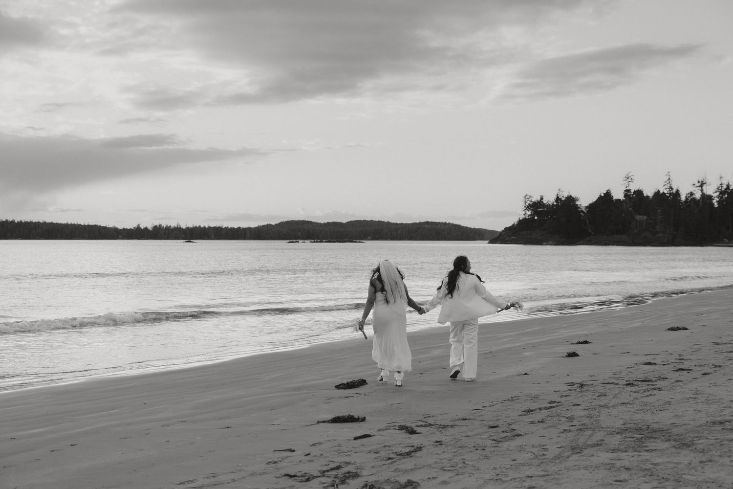 Two people in white outfits hold hands while walking along a serene beach at sunset.
