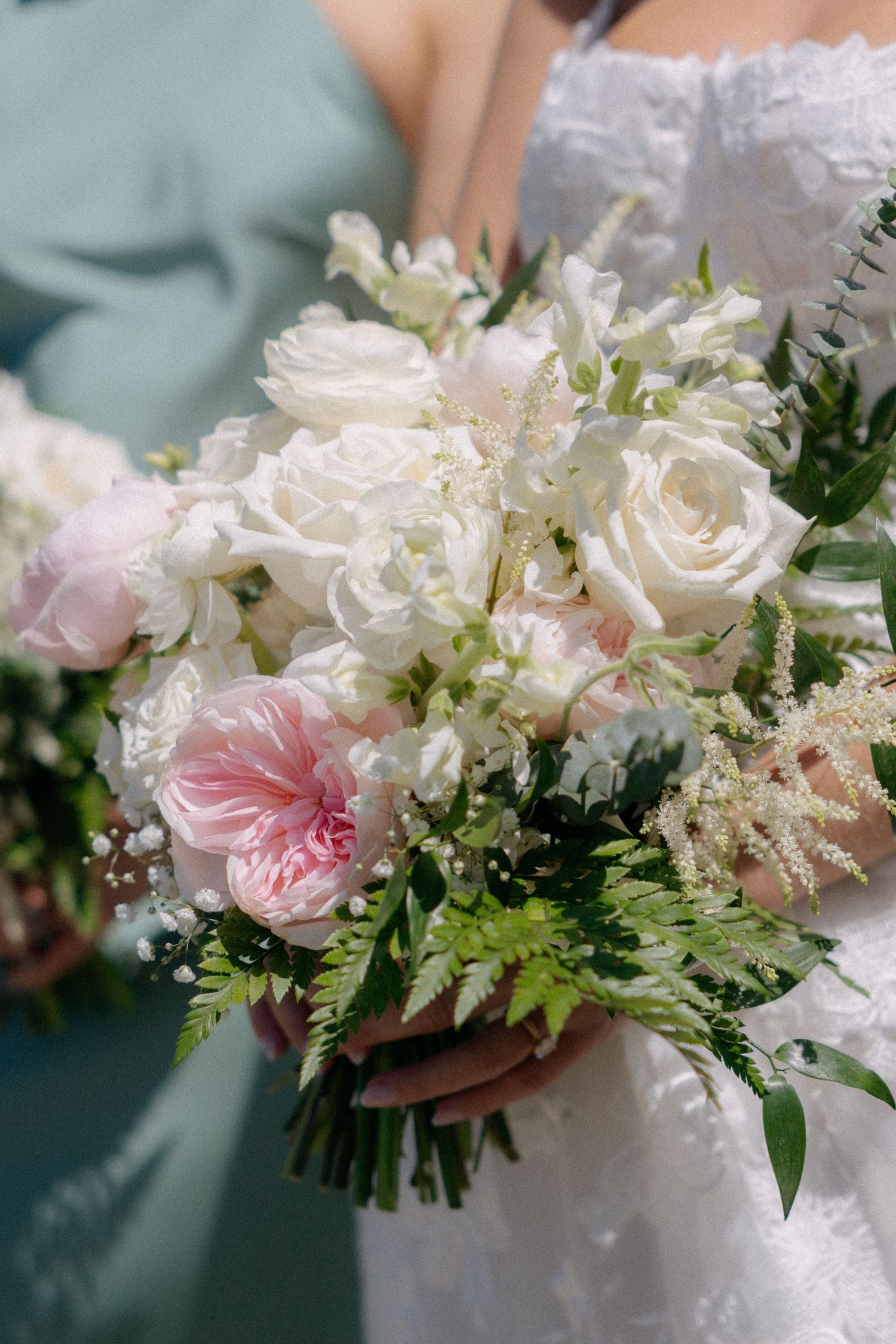 Bride holding elegant wedding bouquet of white roses, pink peonies, and green ferns in soft natural light.