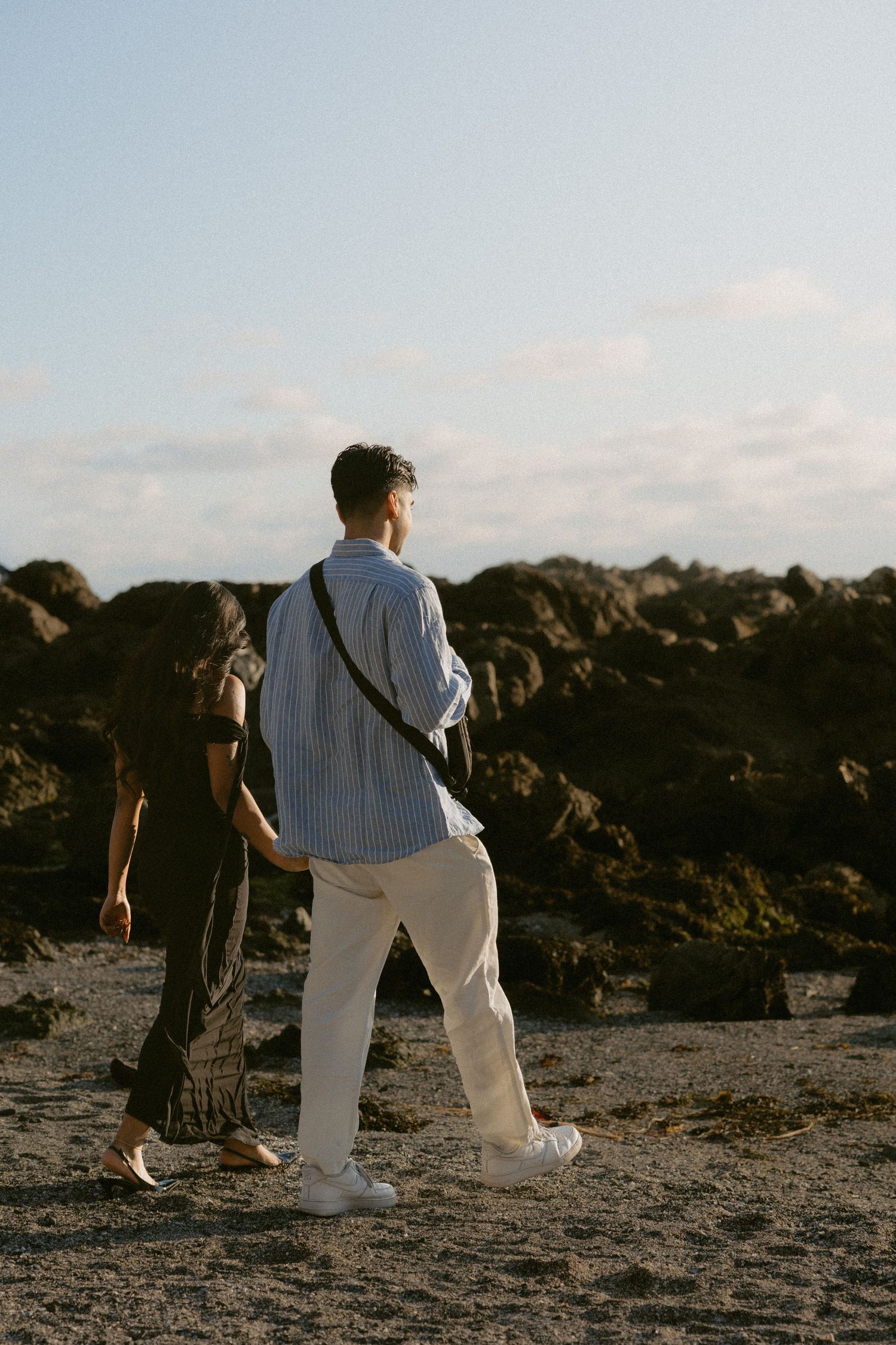 Two people walking on a rocky beach, man in striped shirt and white pants with crossbody bag, woman in dark floral dress.