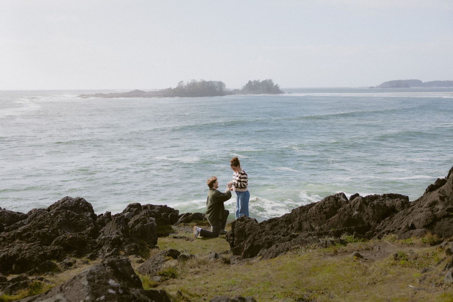 Couple getting engaged on rocky coastal cliffs with ocean waves and misty islands in the background.