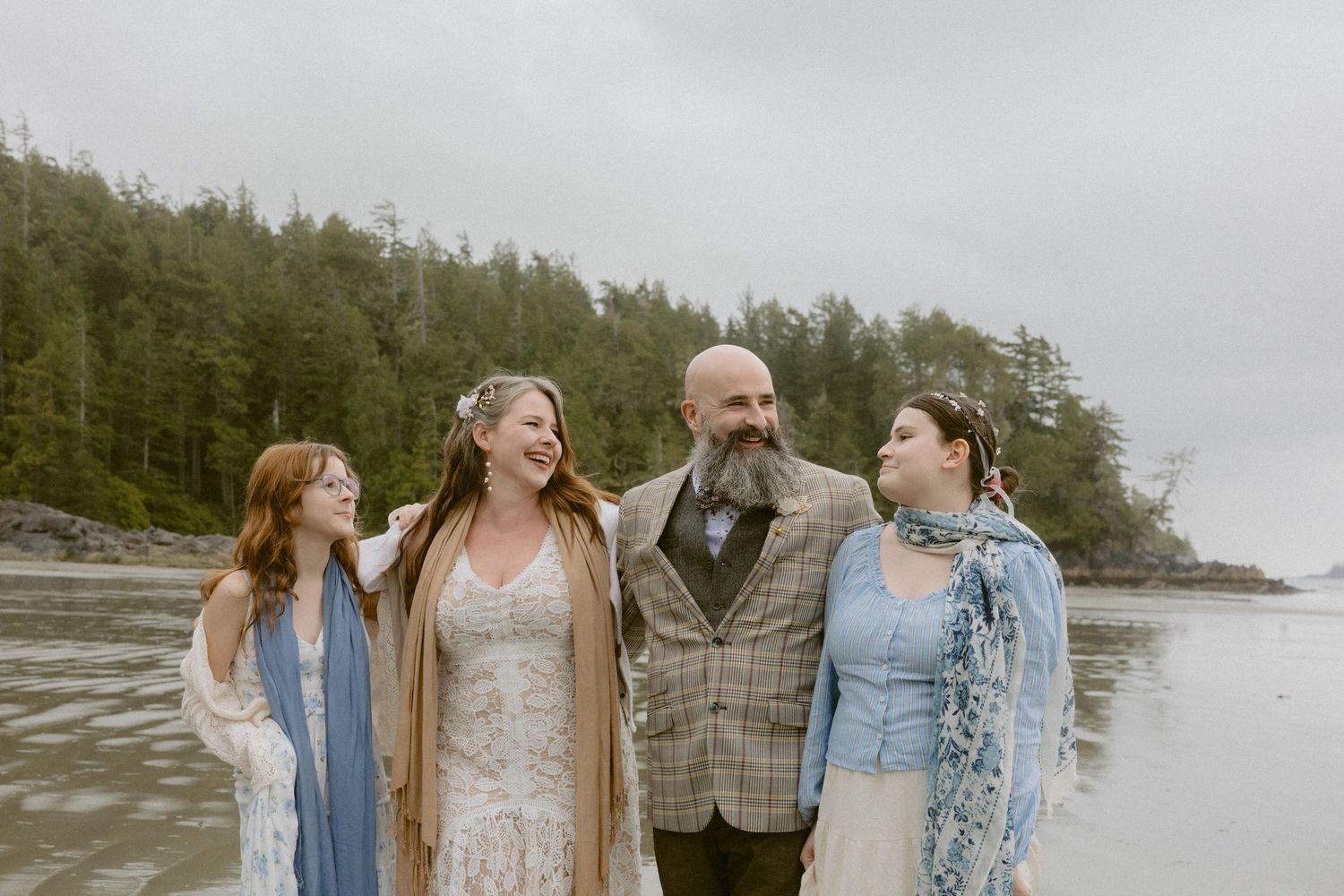 A family group poses together on a moody Pacific Northwest beach with forested cliffs behind them.