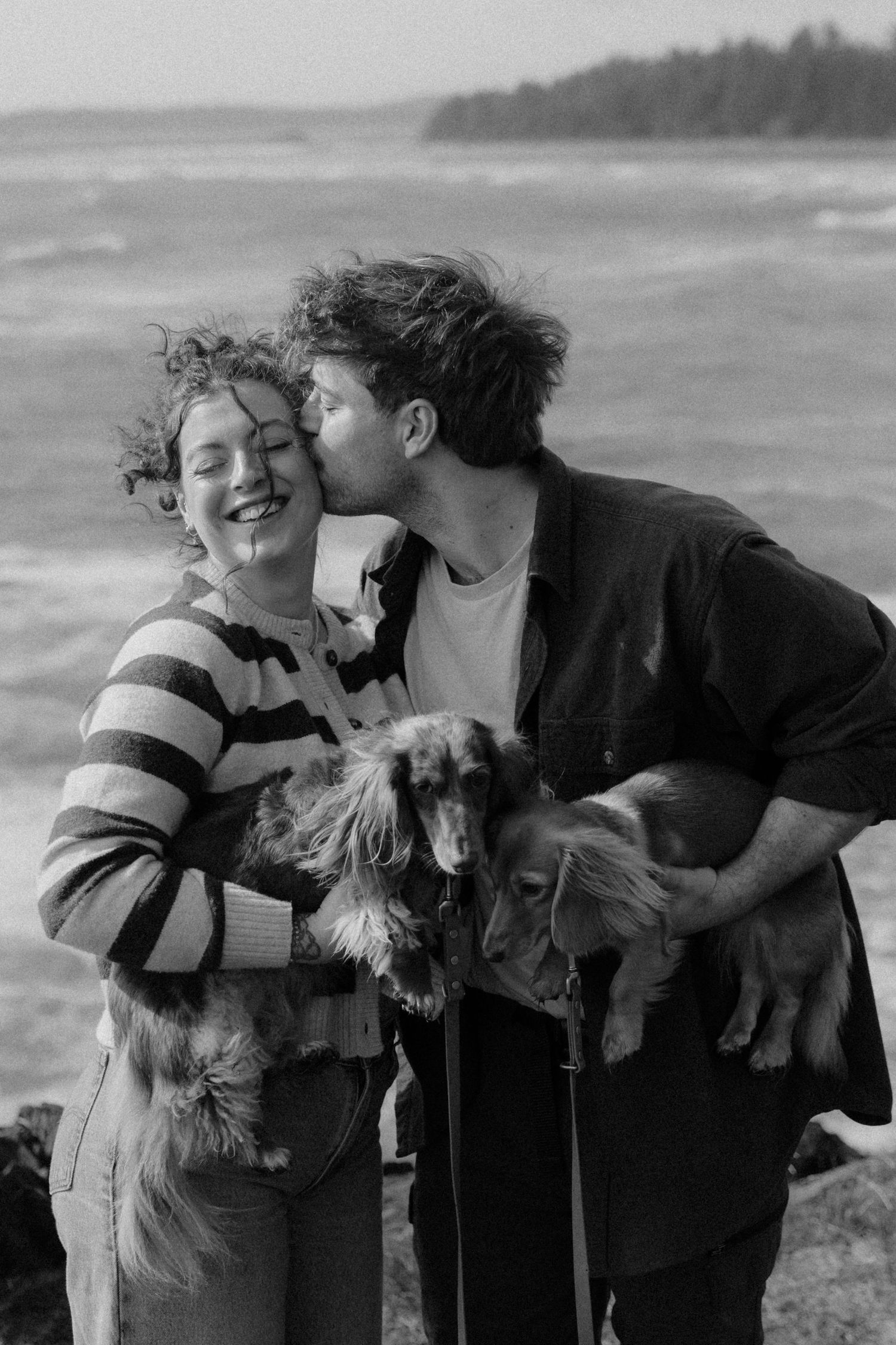 Couple kissing and laughing while holding two long-haired dachshund dogs by the ocean.
