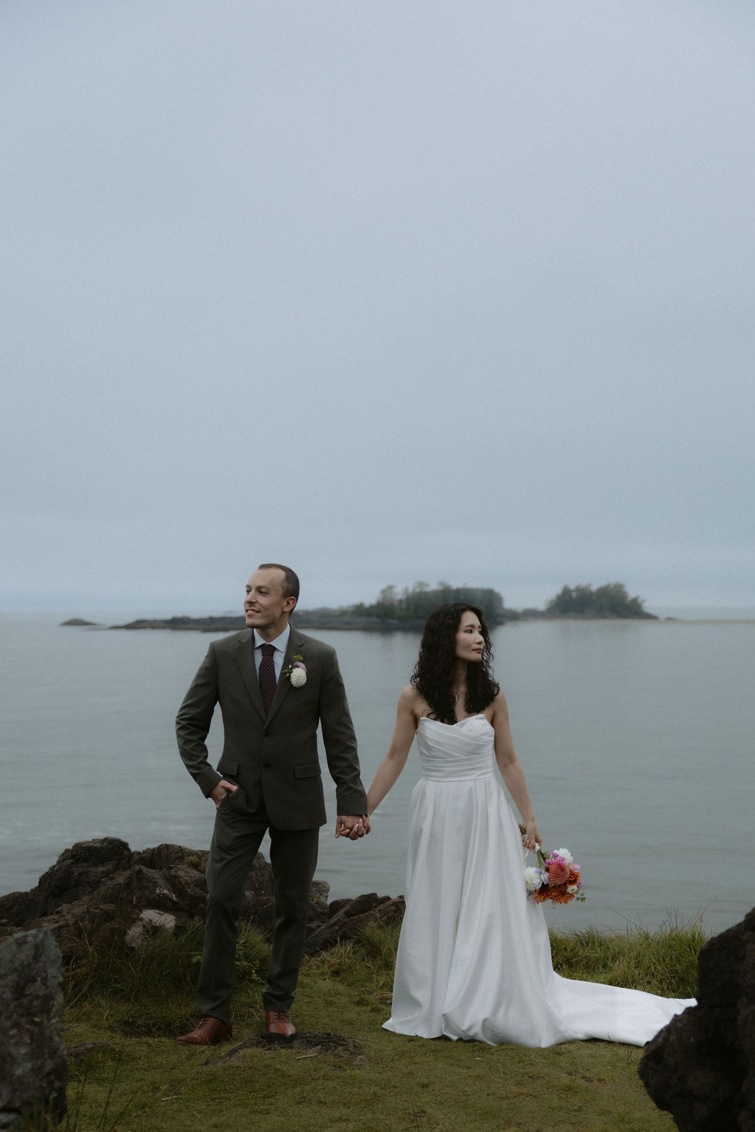 Bride in white gown and groom in olive suit hold hands on rocky coastal cliffside with misty ocean and island backdrop.