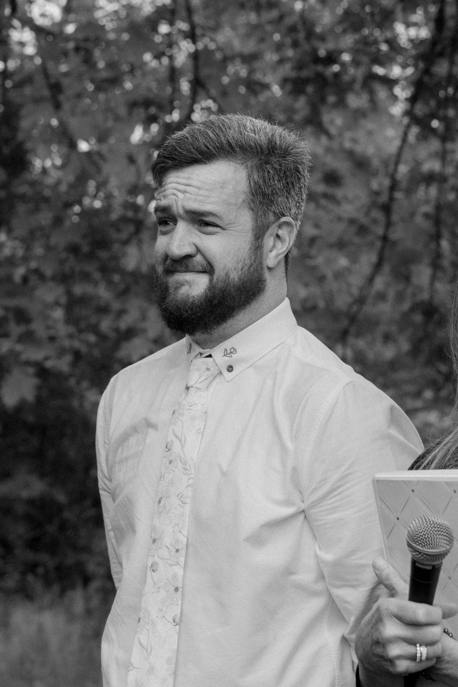 Black and white portrait of bearded man in white shirt and floral tie holding microphone outdoors.