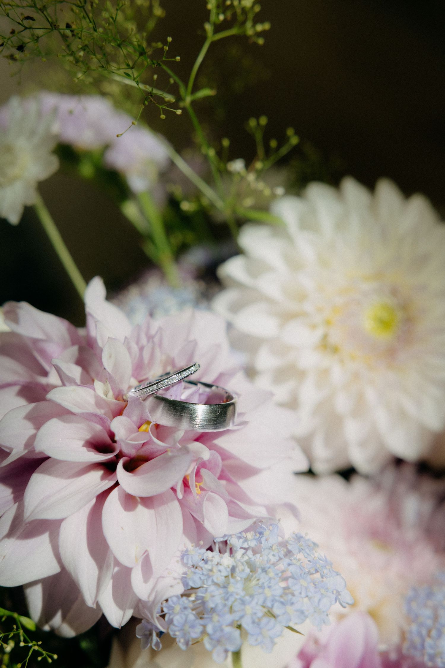 Wedding rings resting on pink dahlia flowers in a bridal bouquet with white dahlias and blue flowers.