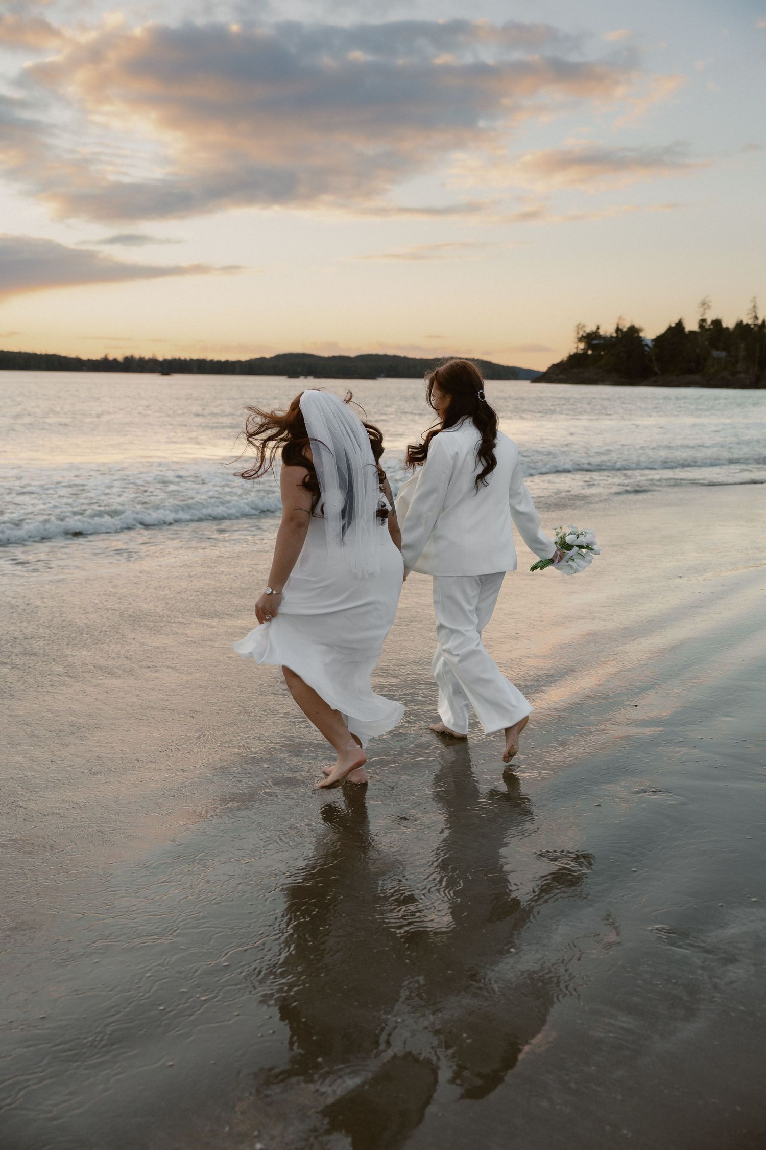 Two people in white clothing walk together through shallow waves on a beach at sunset.
