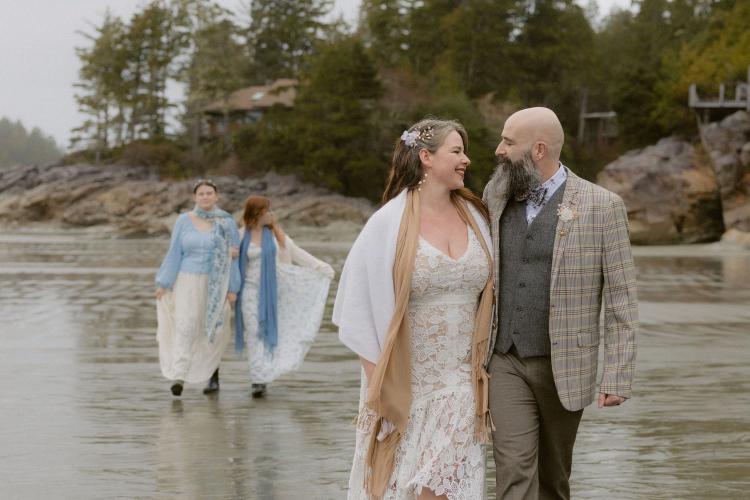 Wedding couple shares a moment on a misty beach with guests walking in the background near rocky cliffs.