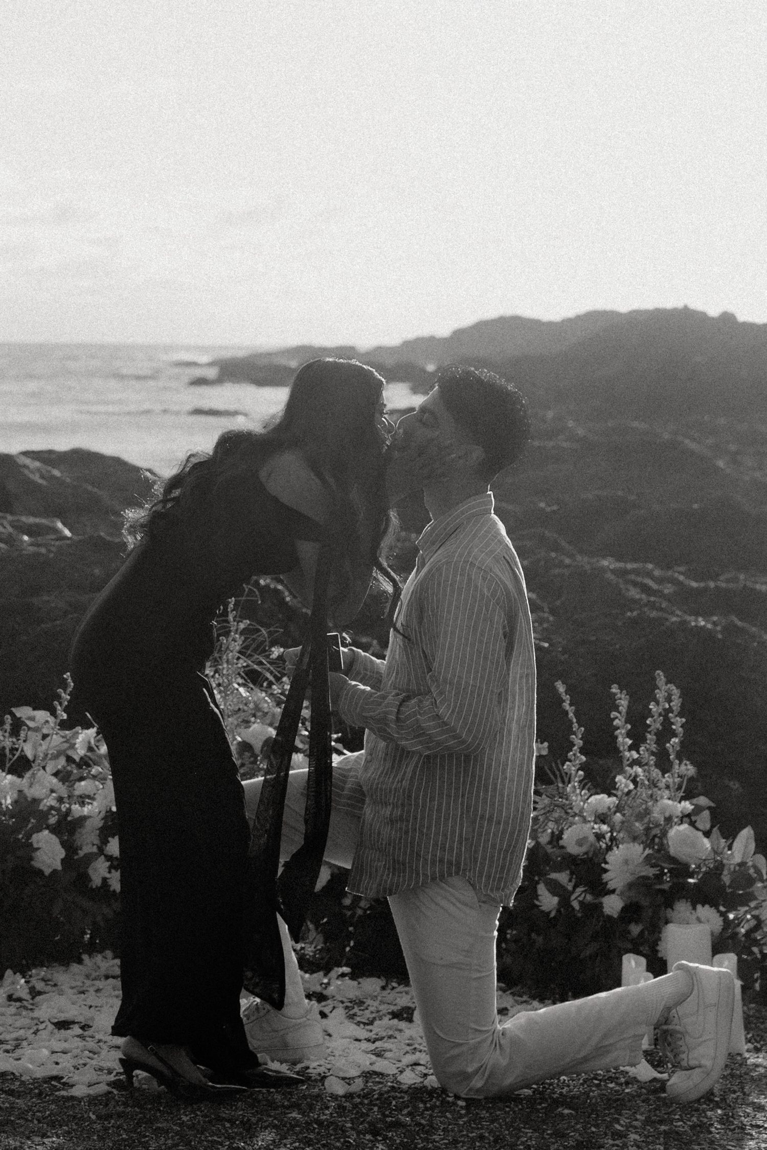 Black and white photo of a man on one knee proposing to a woman by the ocean at dusk.