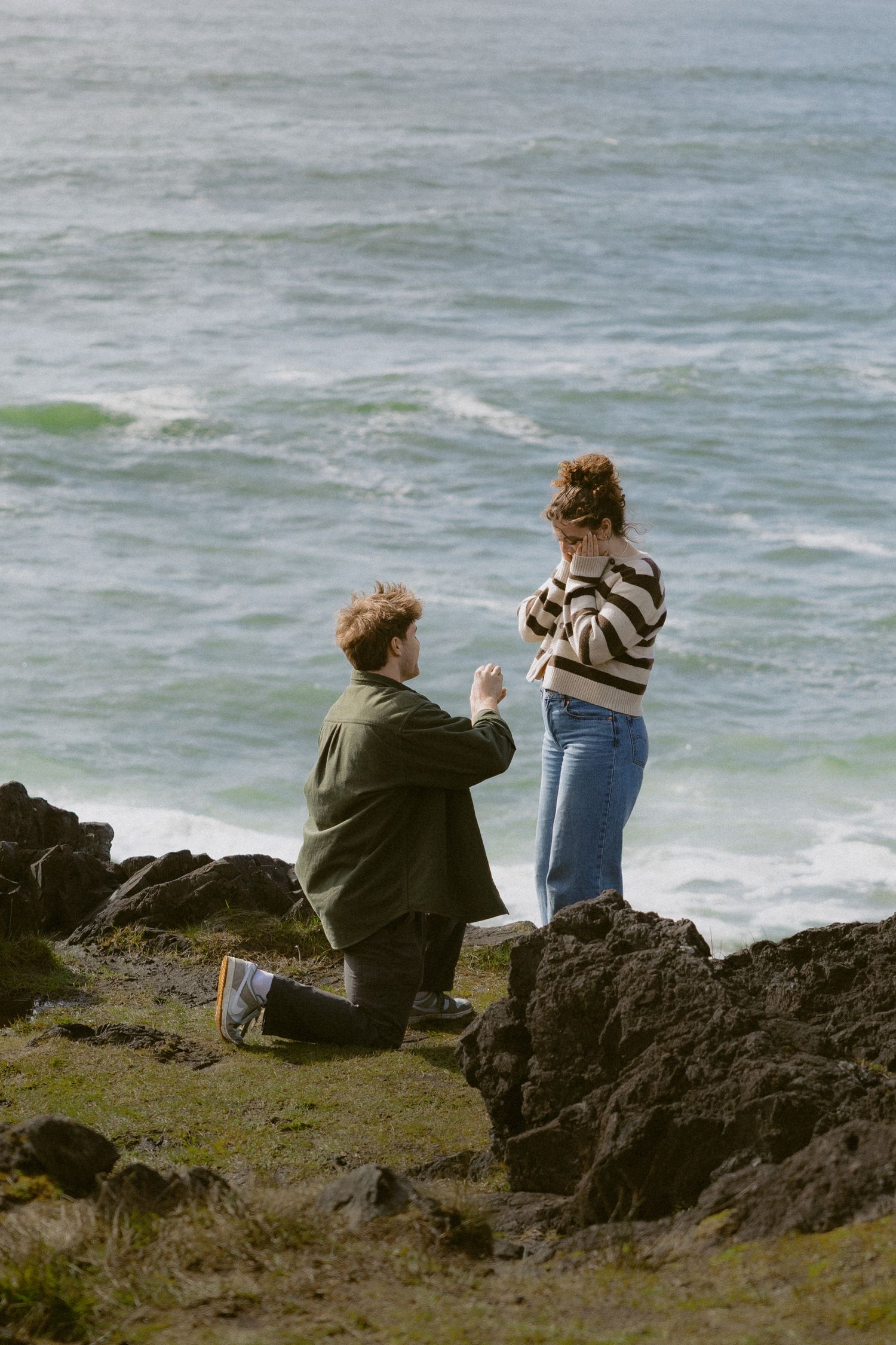 Man proposing on one knee to woman on rocky coastal cliffs with ocean waves in background.