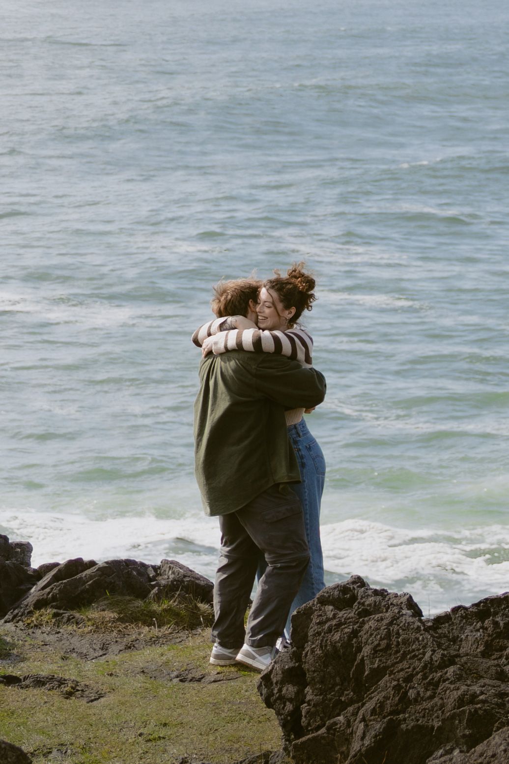 Couple embracing romantically on rocky coastal cliffs with turquoise ocean waves in the background.